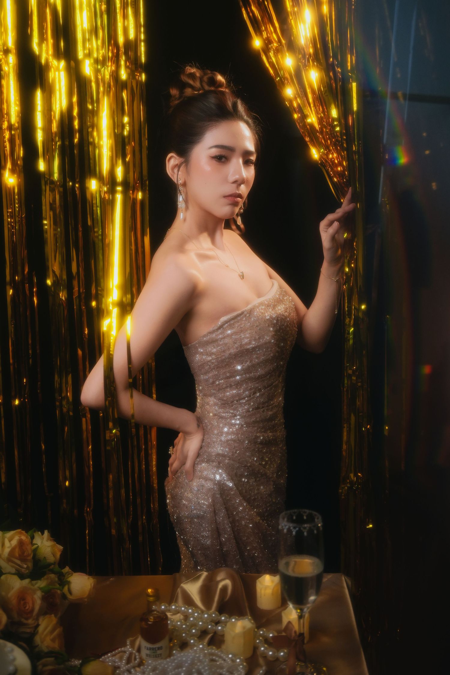 A glamorous figure poses in a sparkly evening gown against a backdrop of shimmering gold streamers and candlelight.