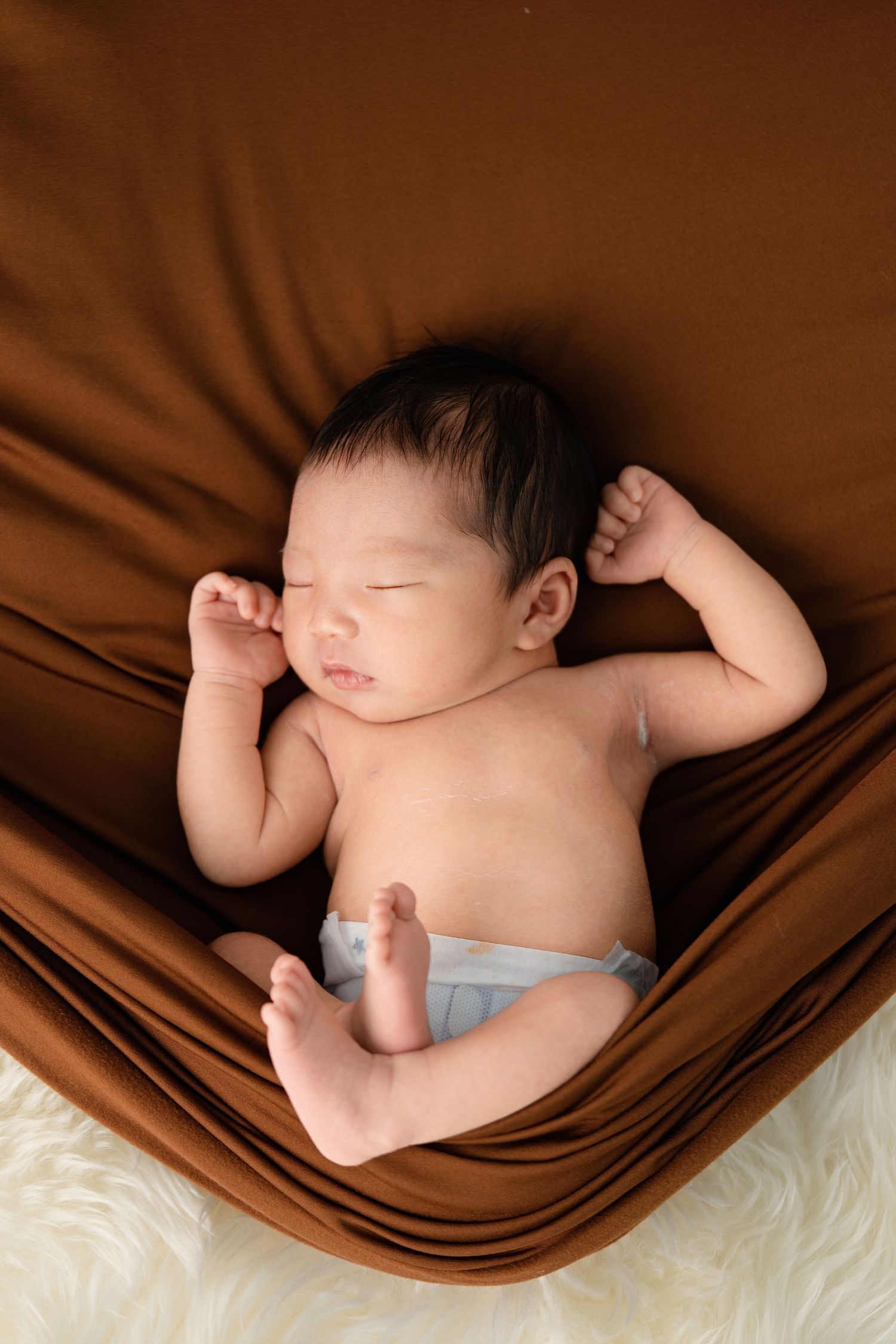 Peaceful newborn baby sleeping on brown fabric wrap, wearing a diaper, with tiny fists raised in a serene portrait.
