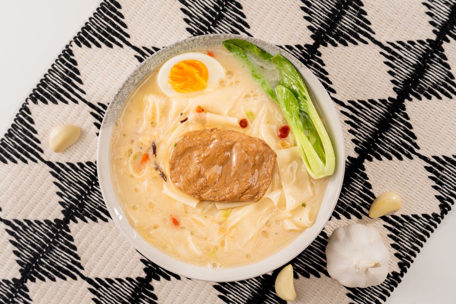 A bowl of creamy ramen with noodles, soft-boiled egg, bok choy, and miso paste on a checkered cloth.