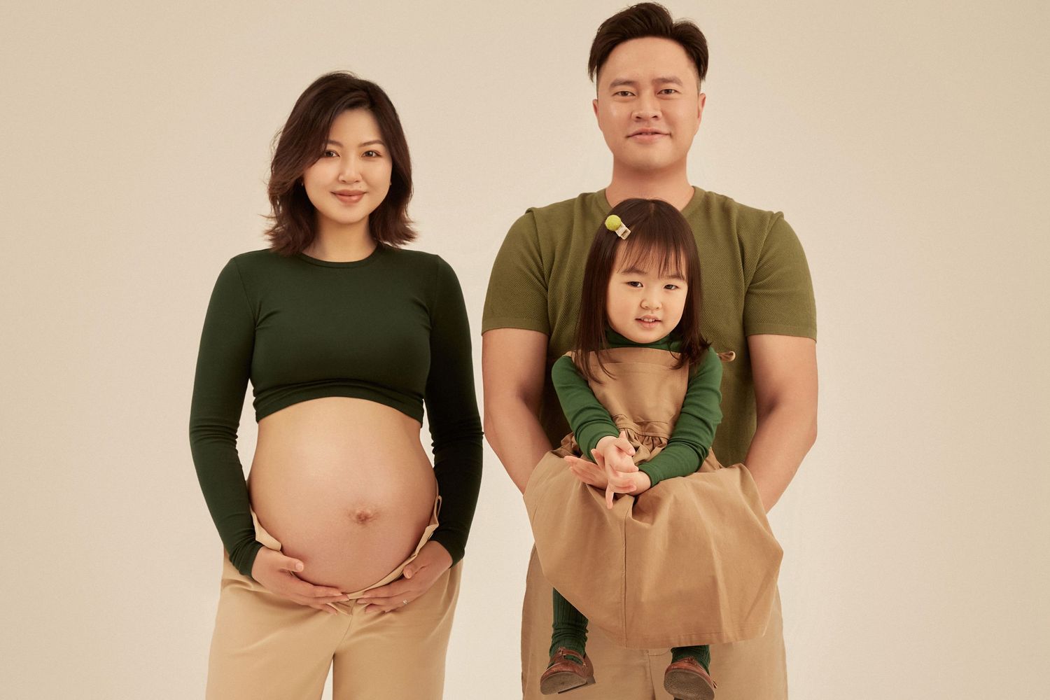 Pregnant Asian woman with family, husband holding toddler daughter, maternity photo shoot on beige background.
