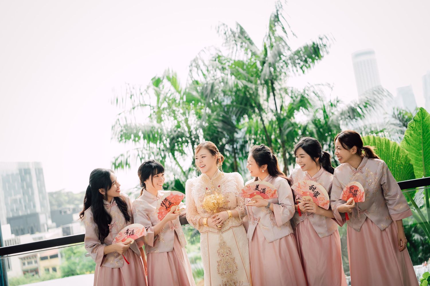 Bridesmaids in soft pink dresses gather outdoors at a garden reception venue with lush greenery and city views.