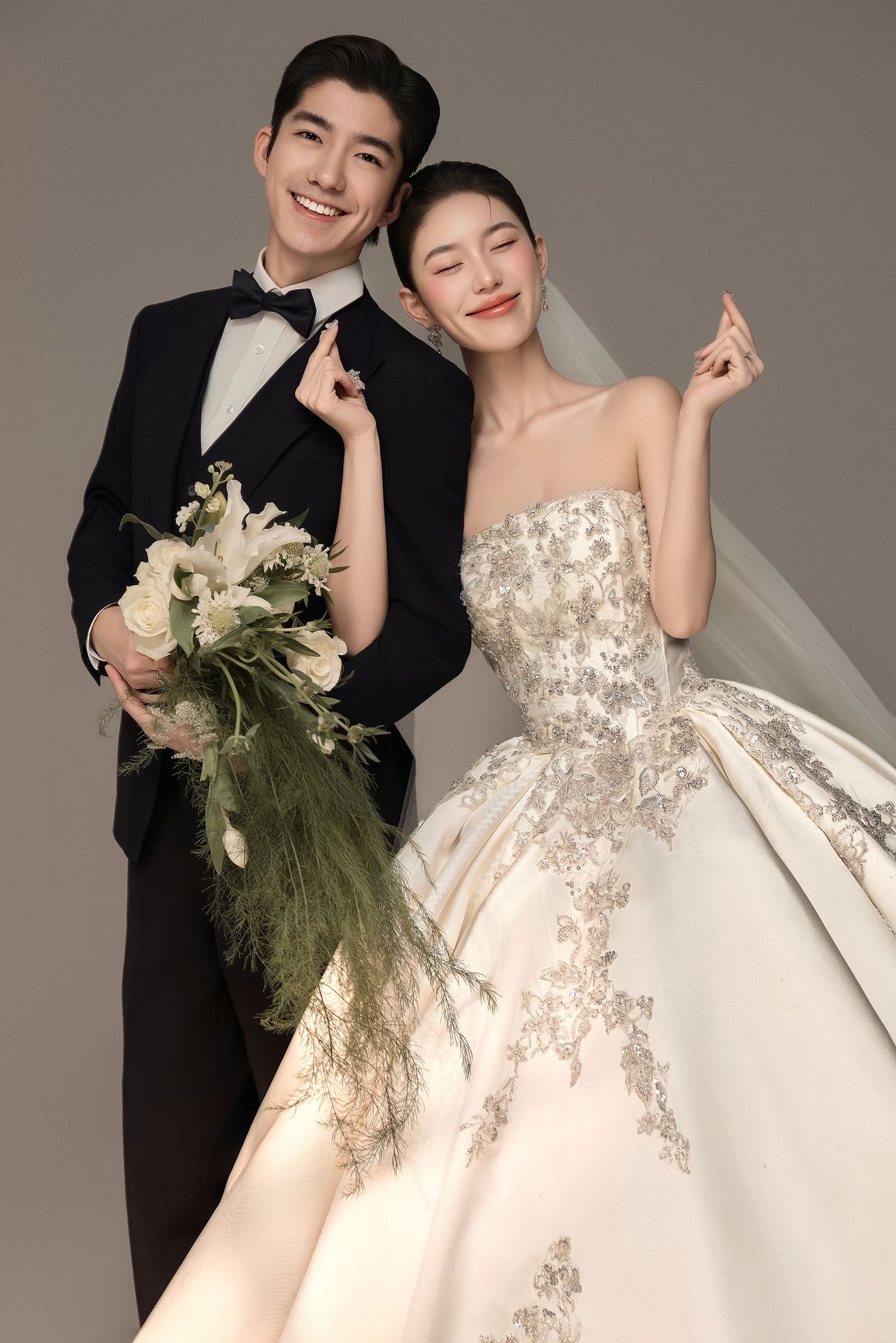 Elegant couple in wedding attire, groom in black tuxedo, bride in embellished ball gown holding floral bouquet.