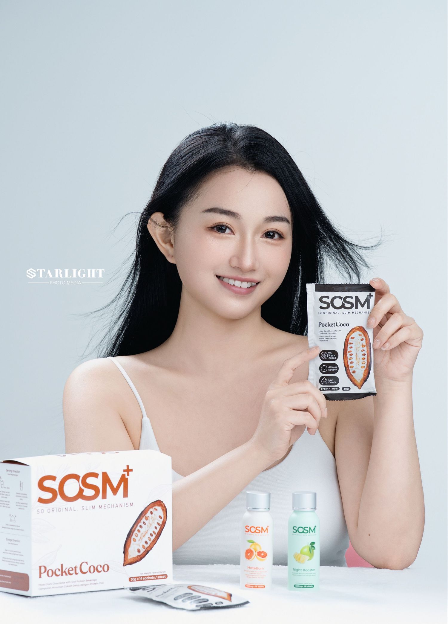 Young woman smiling and holding SOSM PocketCoco product box, with additional SOSM health supplements displayed on white table.