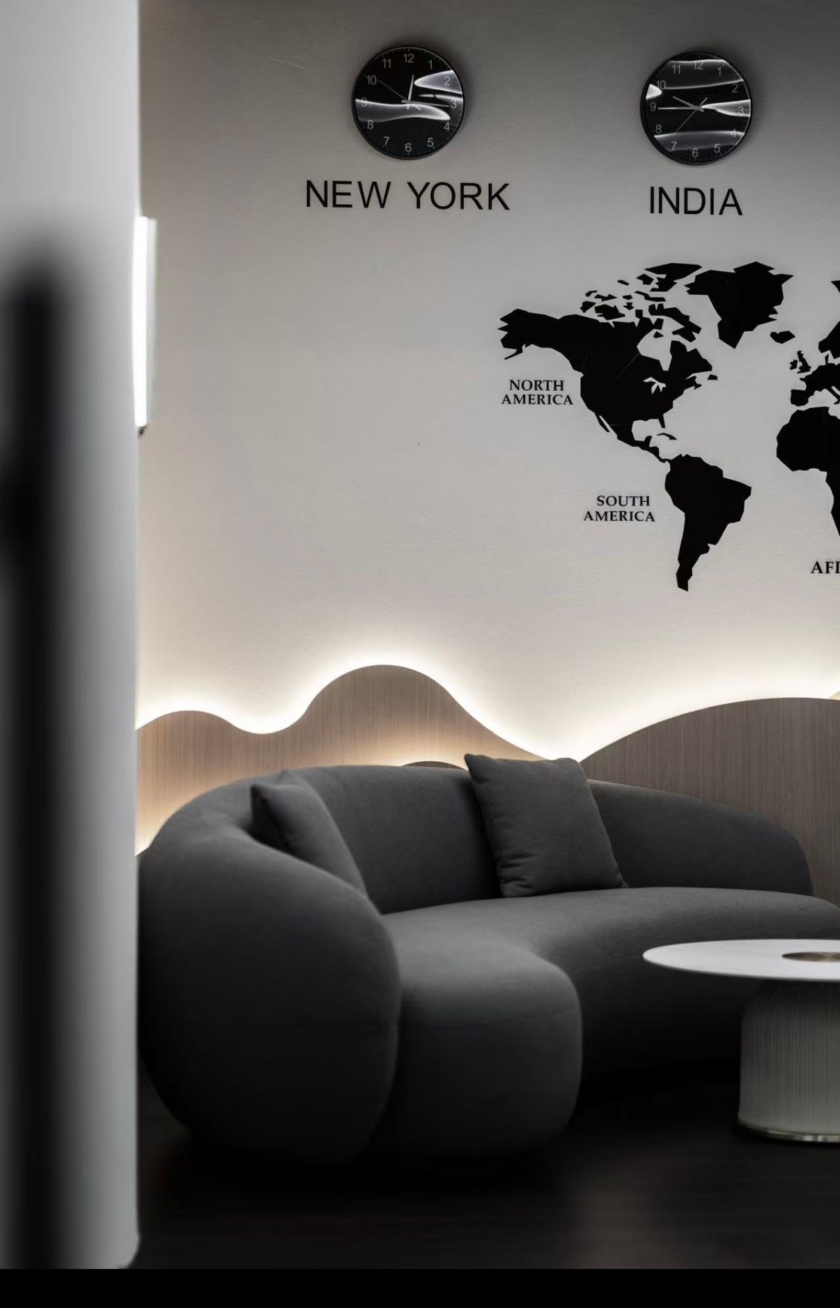 Modern lounge with curved gray sofa, world map wall art, and clocks showing New York and India time zones.