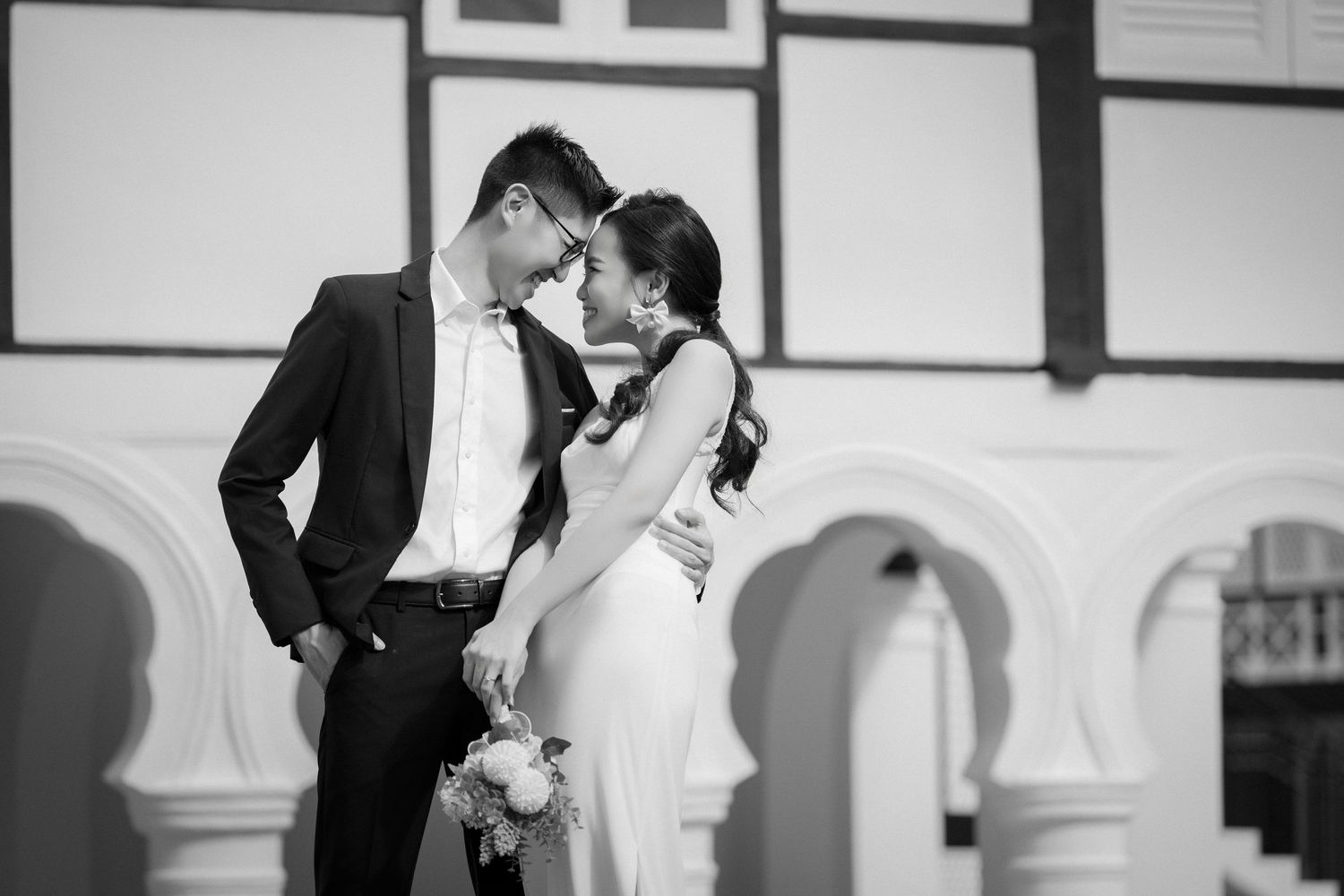 Black and white photo of a couple in wedding attire sharing an intimate moment outside a building with arched columns.