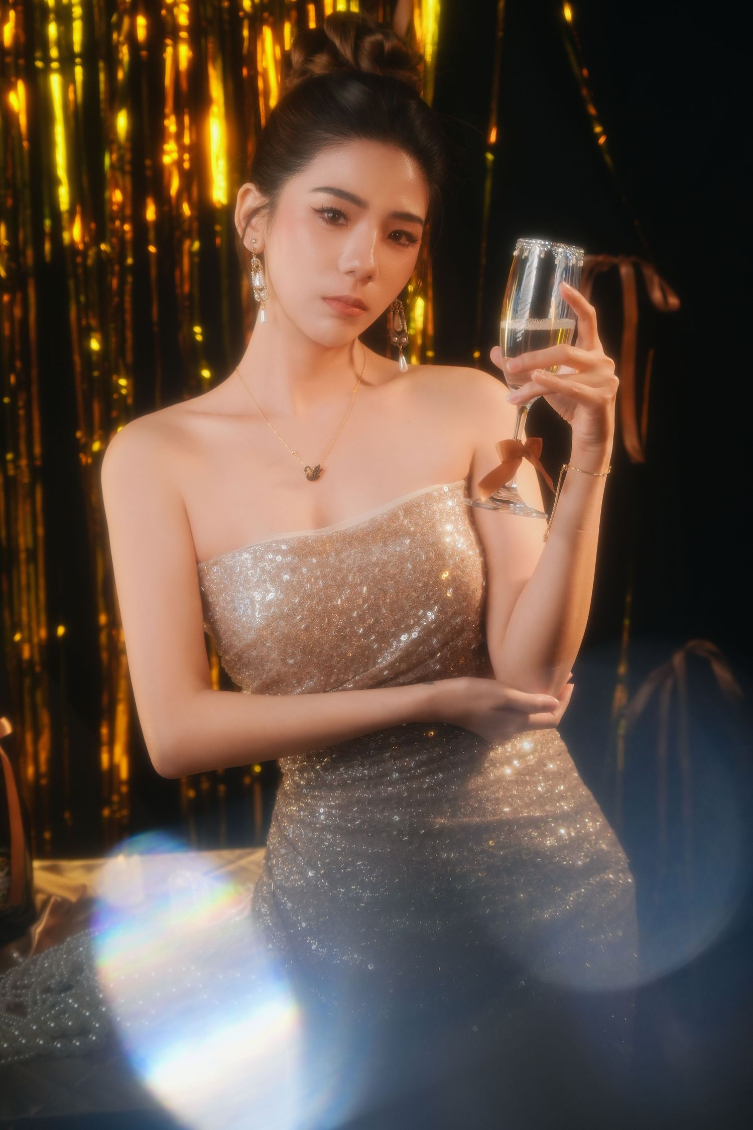 A woman in a gold sequin strapless gown holds a champagne glass against a shimmering gold tinsel backdrop.