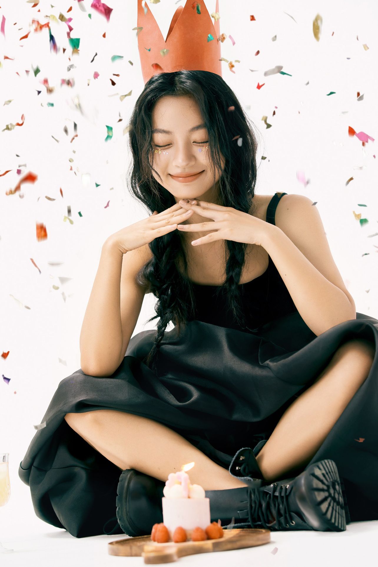 A person in a black dress sits cross-legged with confetti falling while celebrating with a small birthday cake.