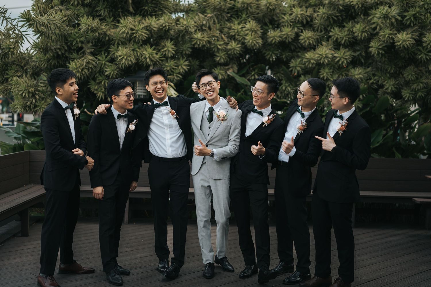 A group of groomsmen in black and grey suits share a candid laughing moment together on a wooden deck.