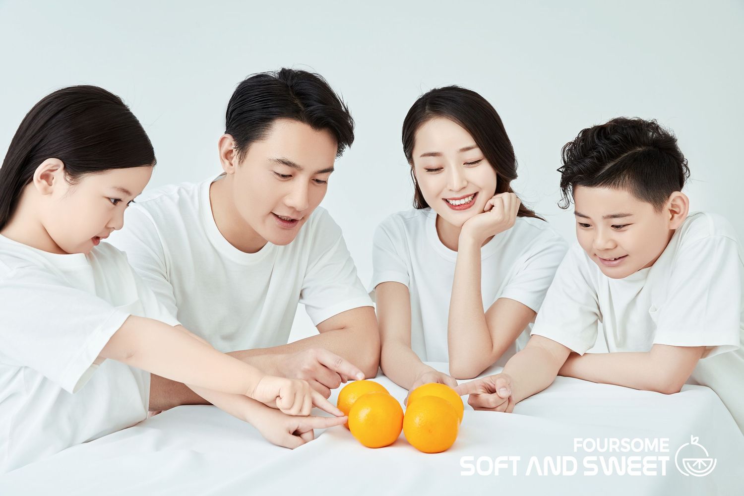 Happy Asian family of four in white shirts reaching for oranges on a white surface, smiling together.