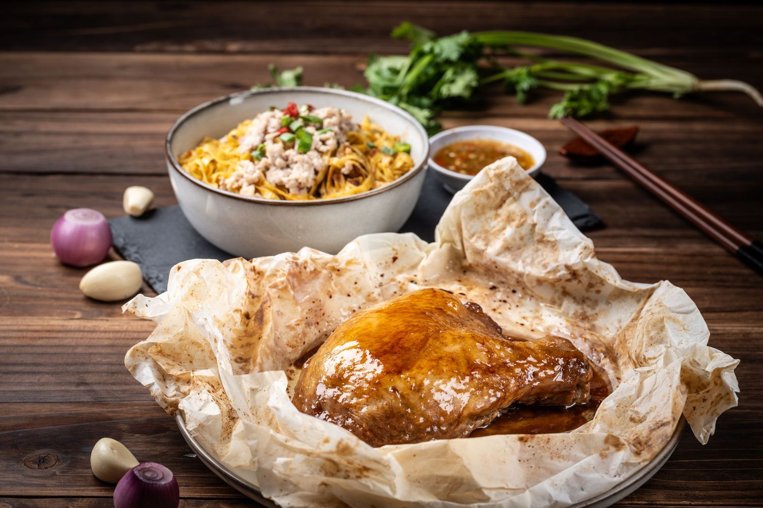 Paper-wrapped roasted chicken served on a plate with a bowl of noodles, garlic, and shallots on a wooden table.