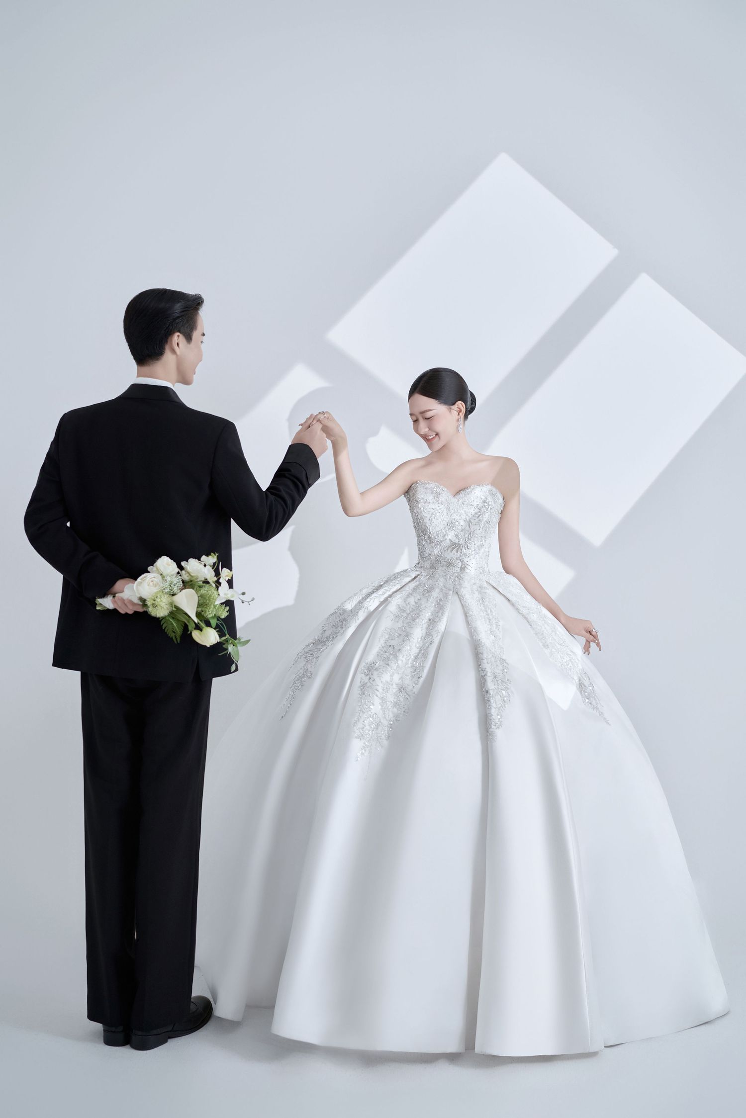 A bride in a white ball gown and groom in black tuxedo hold hands, dancing elegantly in a bright studio setting.