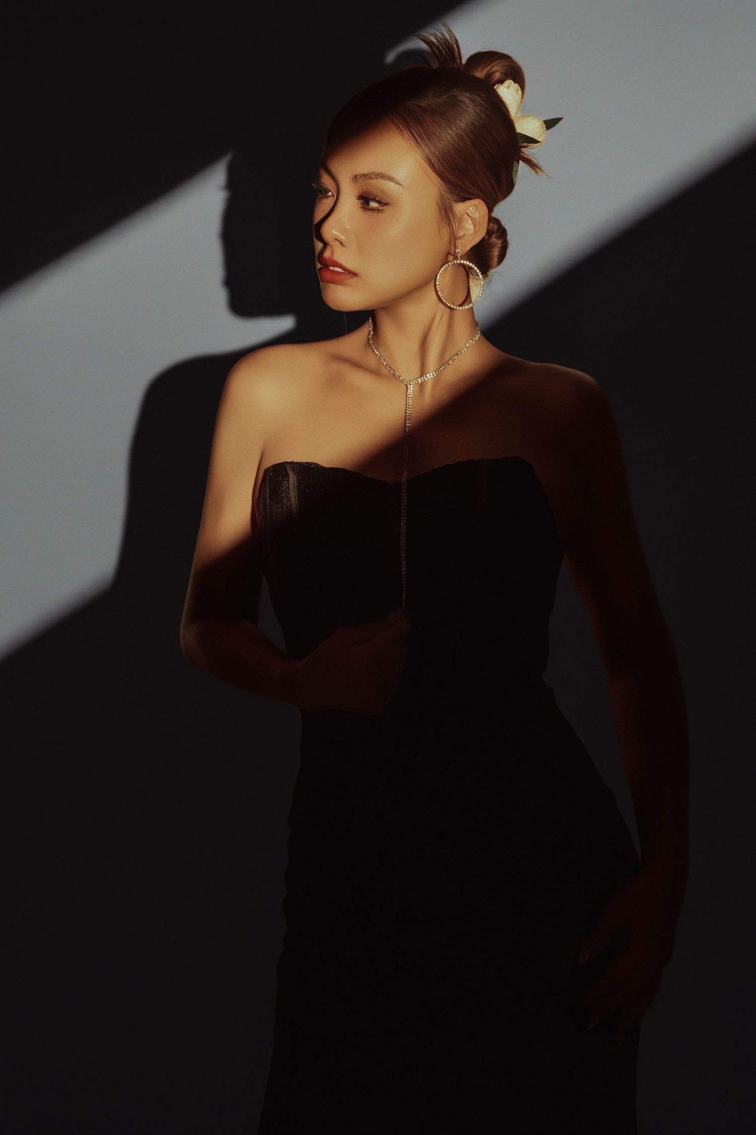 Woman in black strapless dress with gold hoop earrings and hair accessory posing in dramatic shadow lighting.