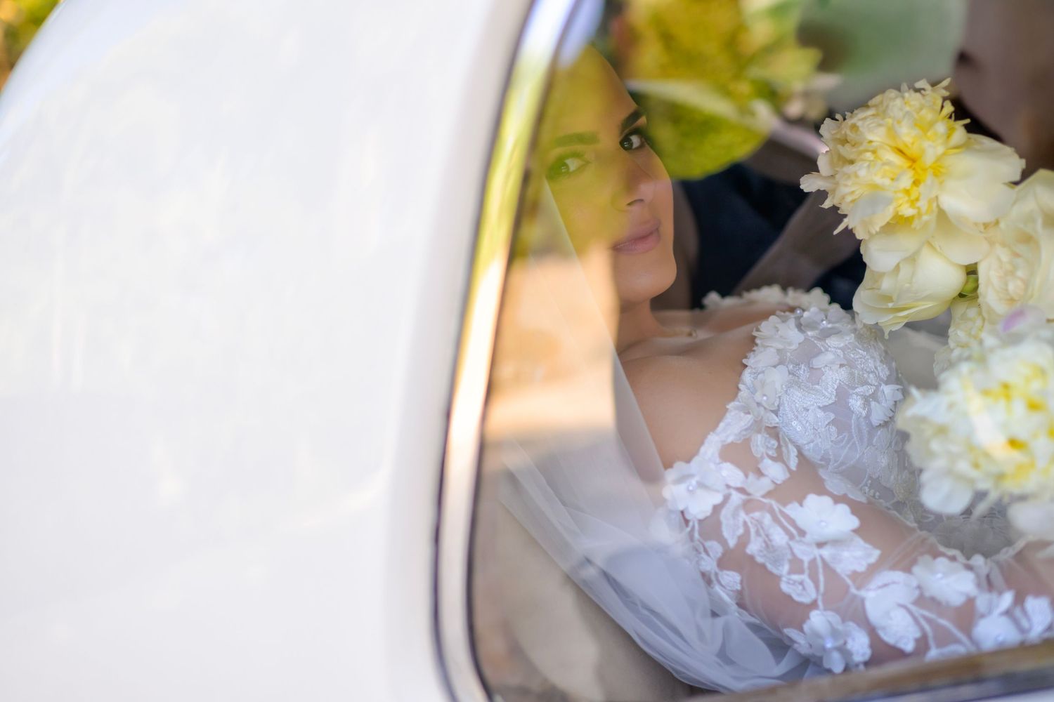 Bride captured through the car window with soft light and yellow florals framing the moment.
