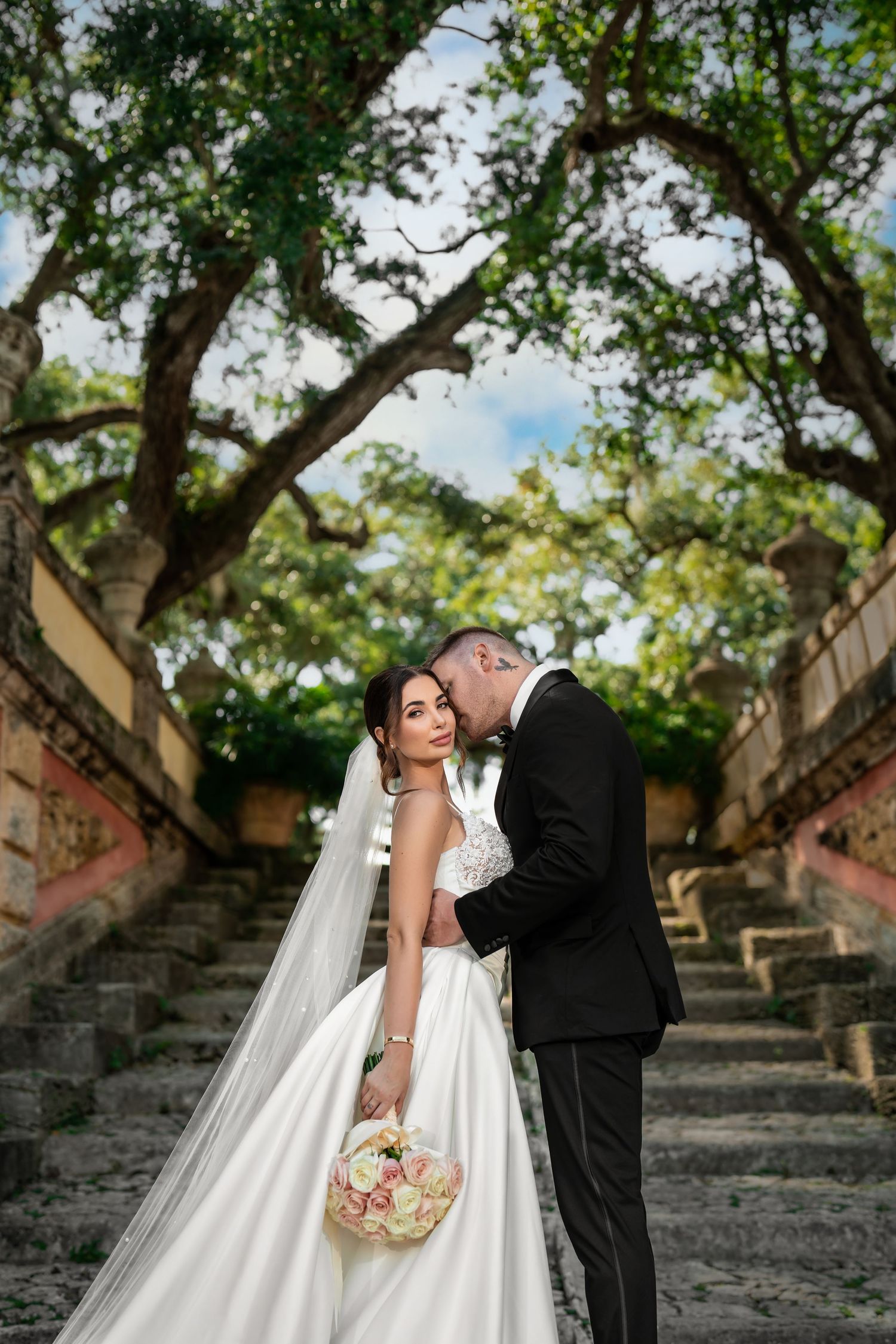 Bride and groom kissing on stone steps surrounded by trees—romantic outdoor wedding portrait.