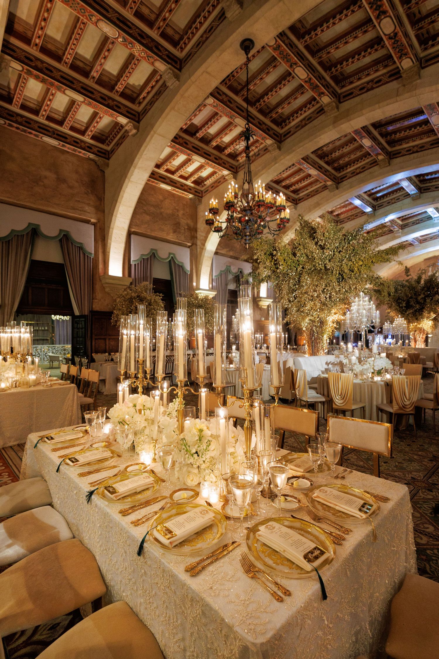 Rows of beautifully set tables under vaulted ceilings, glowing with candles and hanging greenery.