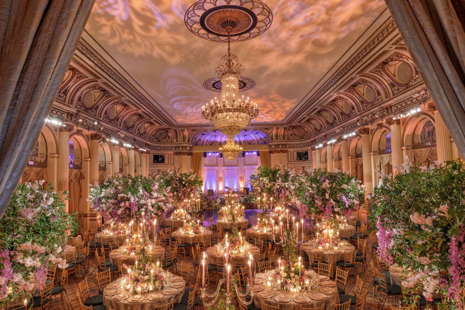 Grand wedding reception at The Plaza in New York—ornate ceiling, lush floral centerpieces, and dramatic uplighting in the ballroom.