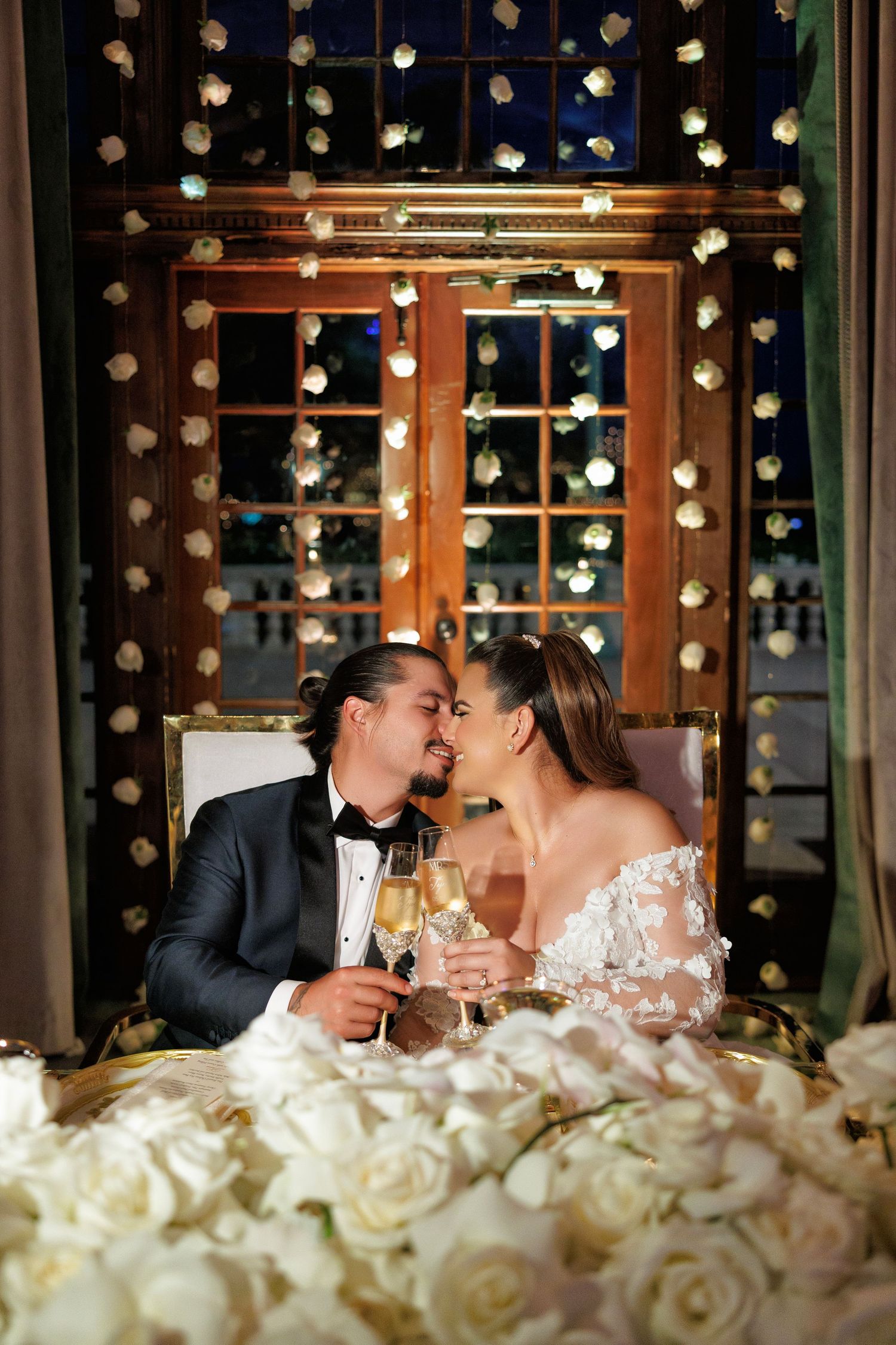 Bride in a glamorous gown sits surrounded by a wall of roses.