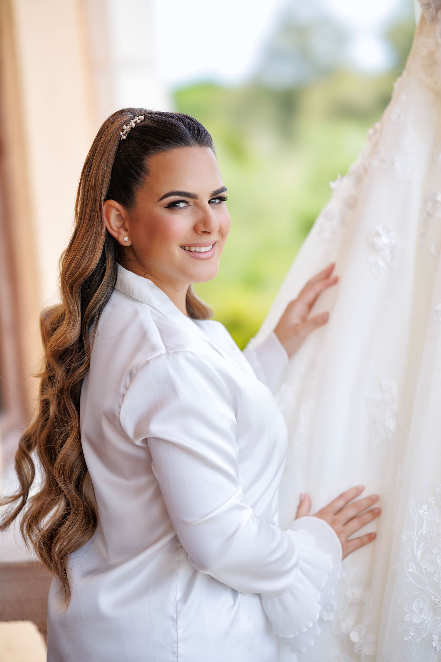 Bride in white robe smiles softly while holding her veil.