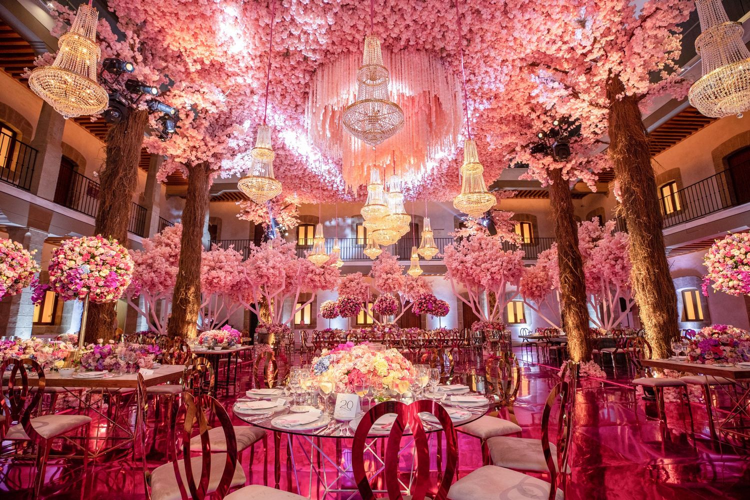 Luxury wedding reception with pink floral ceiling installation, chandeliers, and candlelit tables—lavish ballroom design.
