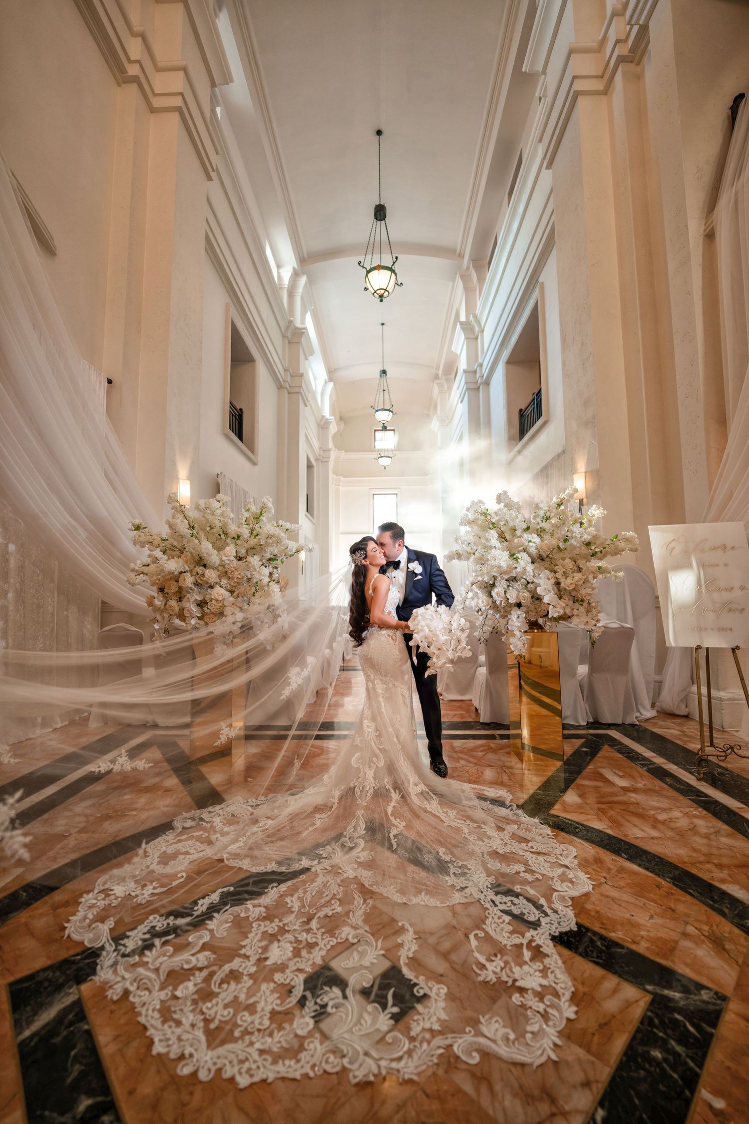 Bride walking through a grand hallway with a cathedral-length train trailing behind—luxury bridal portrait.