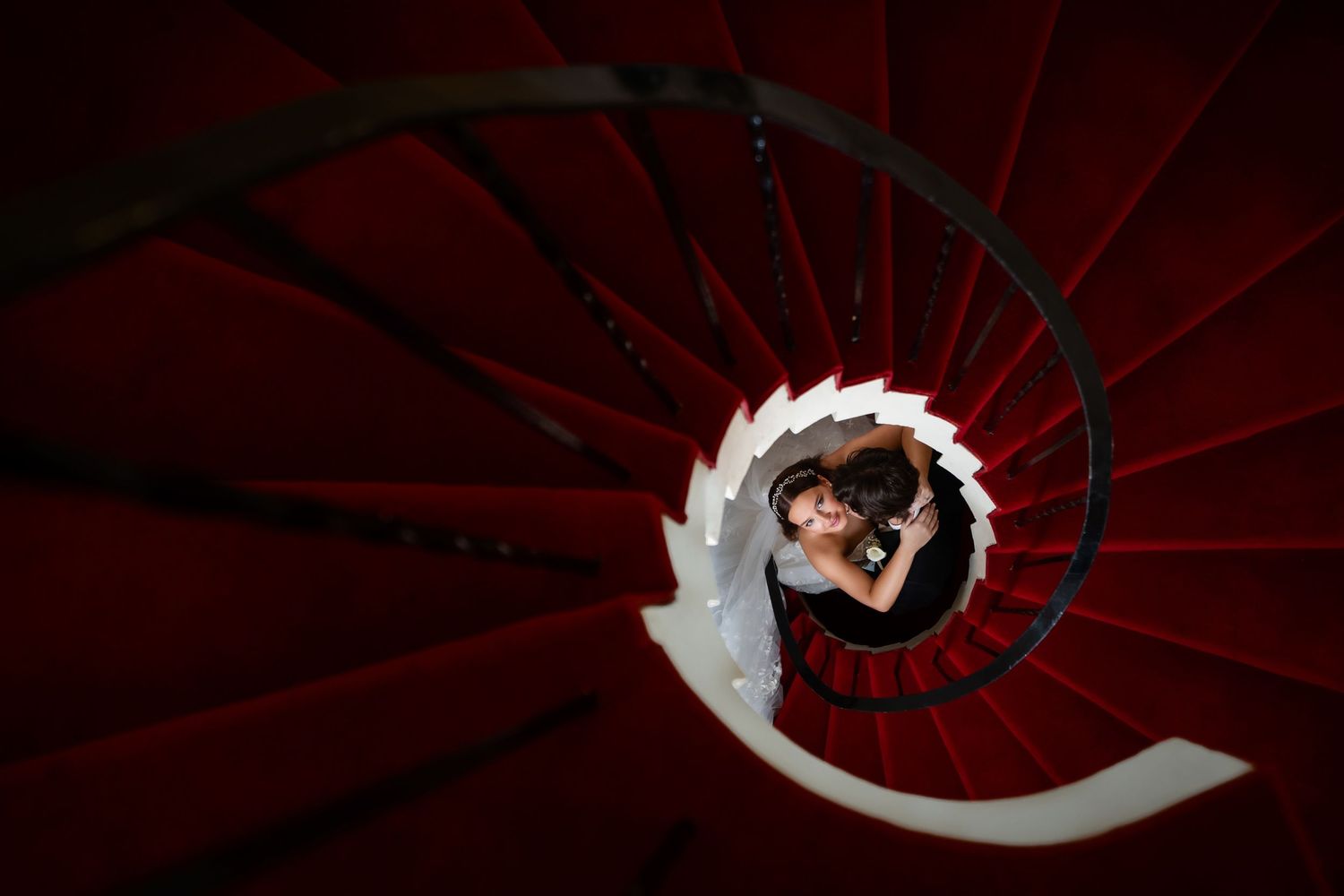 Bride captured from above on a dramatic red spiral staircase—artistic, cinematic wedding portrait.