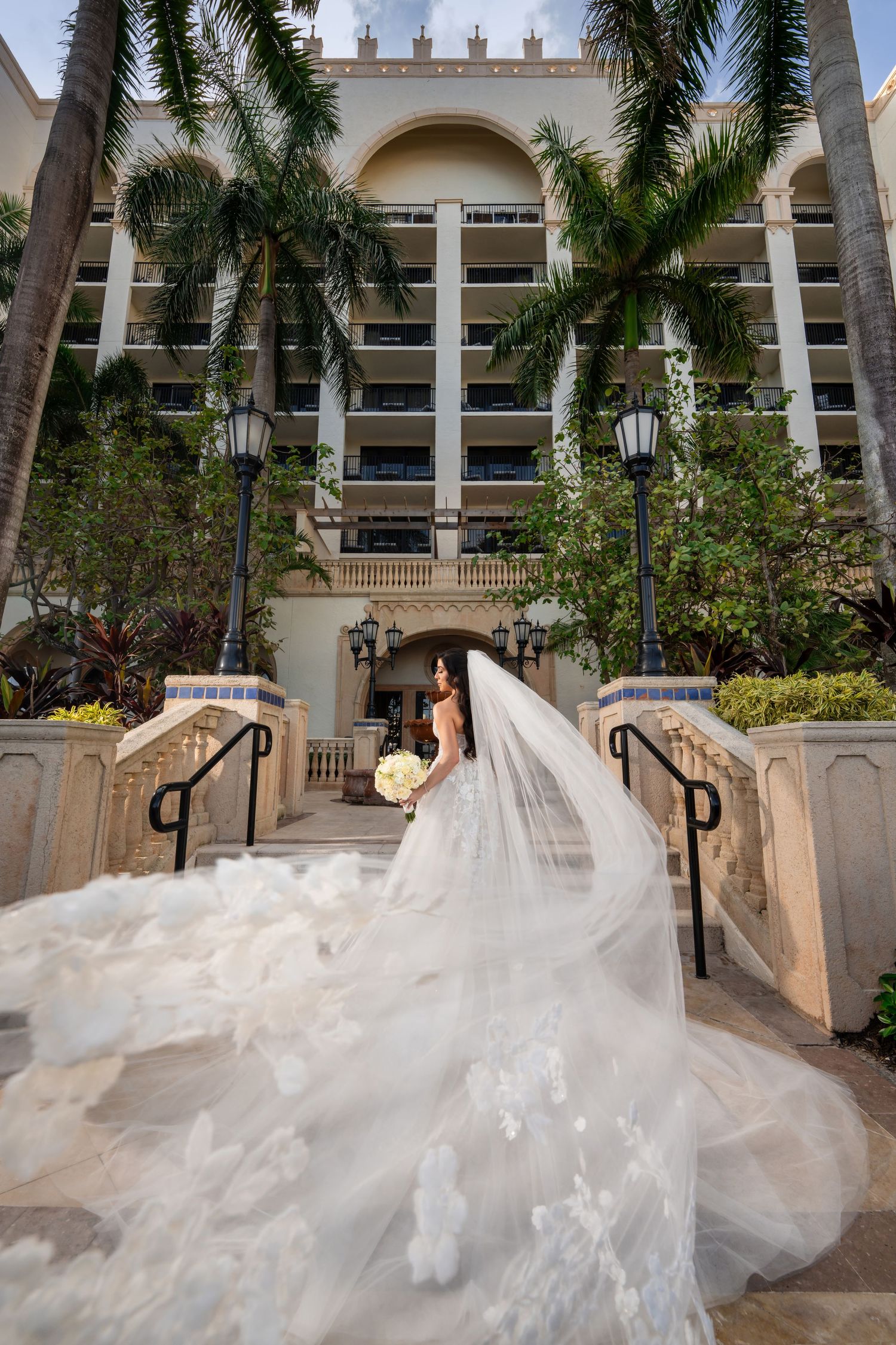 Bride in a voluminous gown posing in a tropical courtyard—luxury wedding portrait with palm trees.