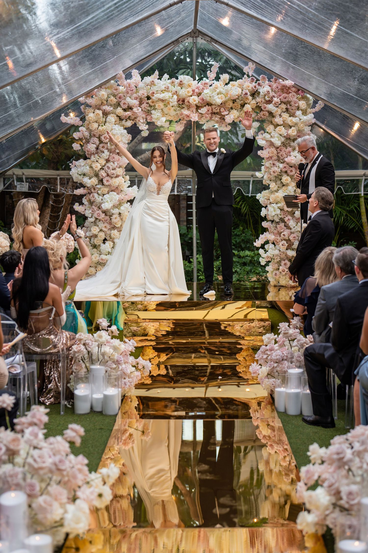 Luxury wedding ceremony inside a glass tent—bride and groom standing beneath a floral arch.