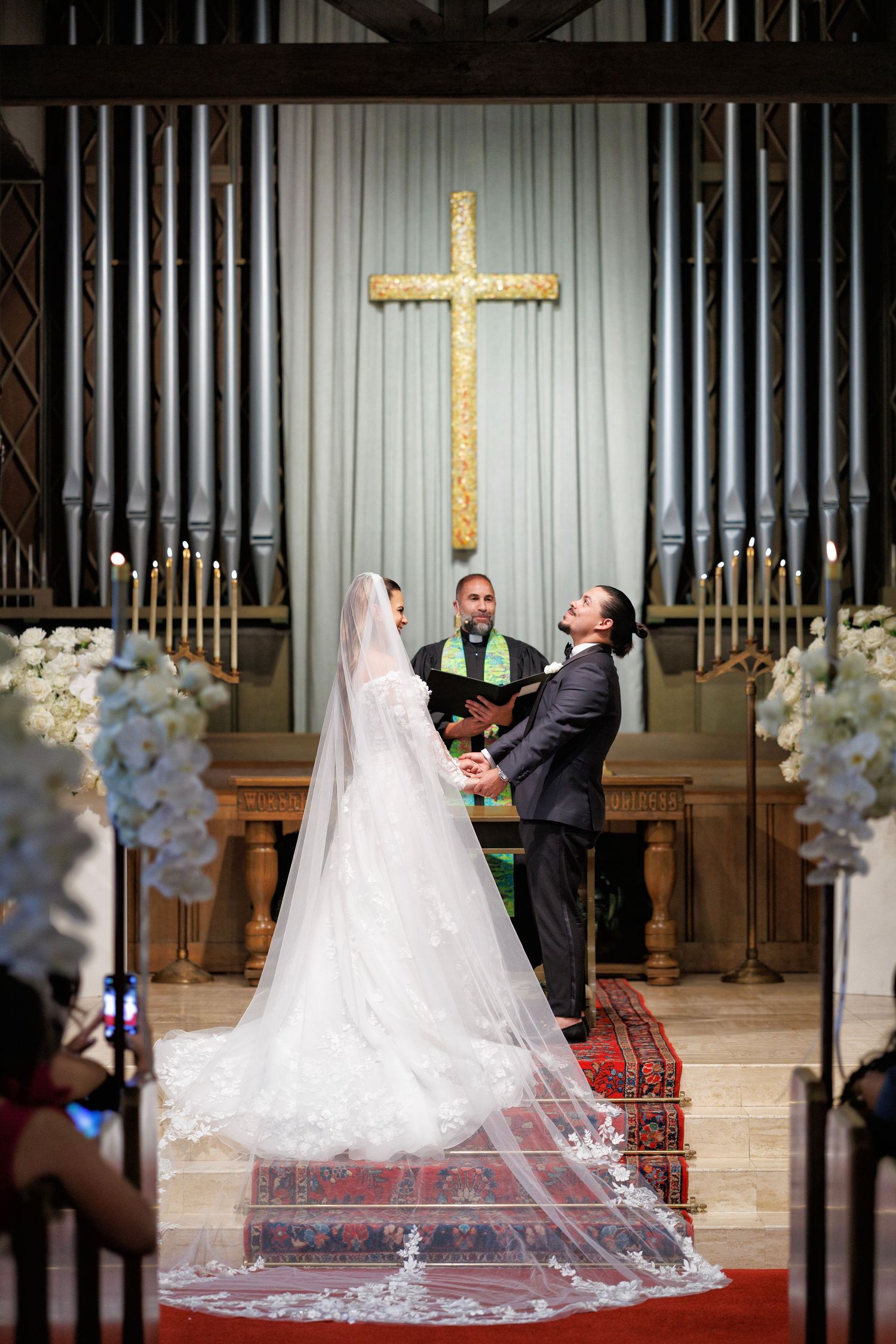 Bride and groom at the altar in a grand church, framed by soaring organ pipes and a golden cross.