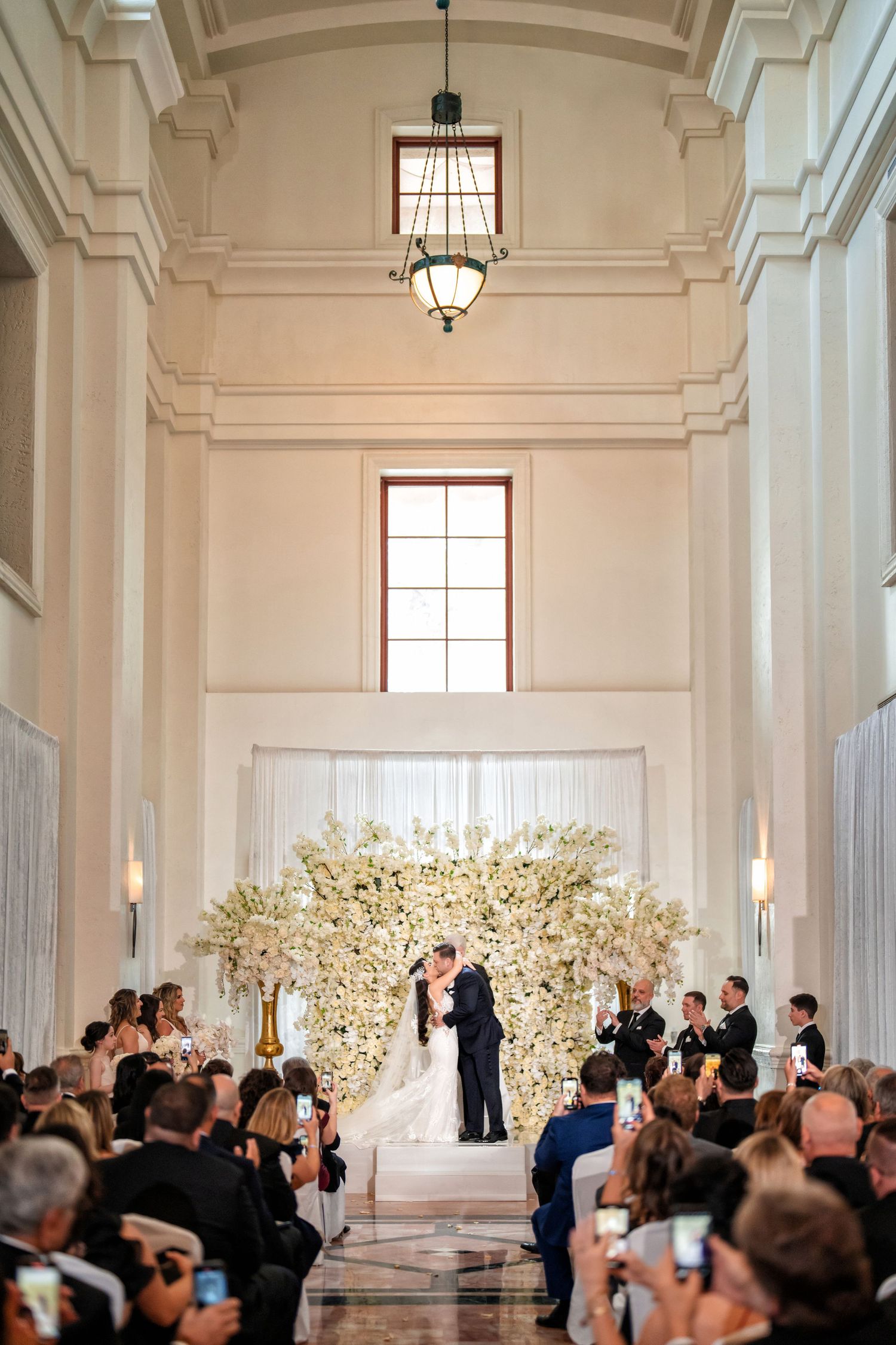 Indoor wedding ceremony with the bride walking down the aisle of a grand white chapel filled with guests.