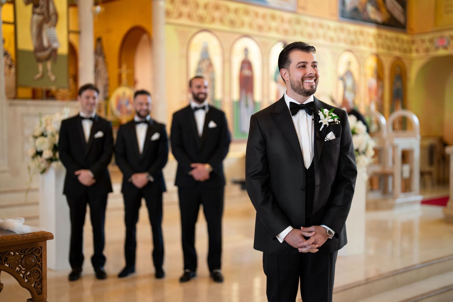 Groom standing with groomsmen in a cathedral—formal wedding portrait in black tuxedos.