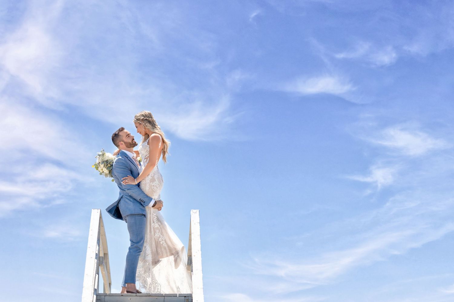Cinematic wedding portrait of a bride and groom kissing on white steps under a bright blue sky.