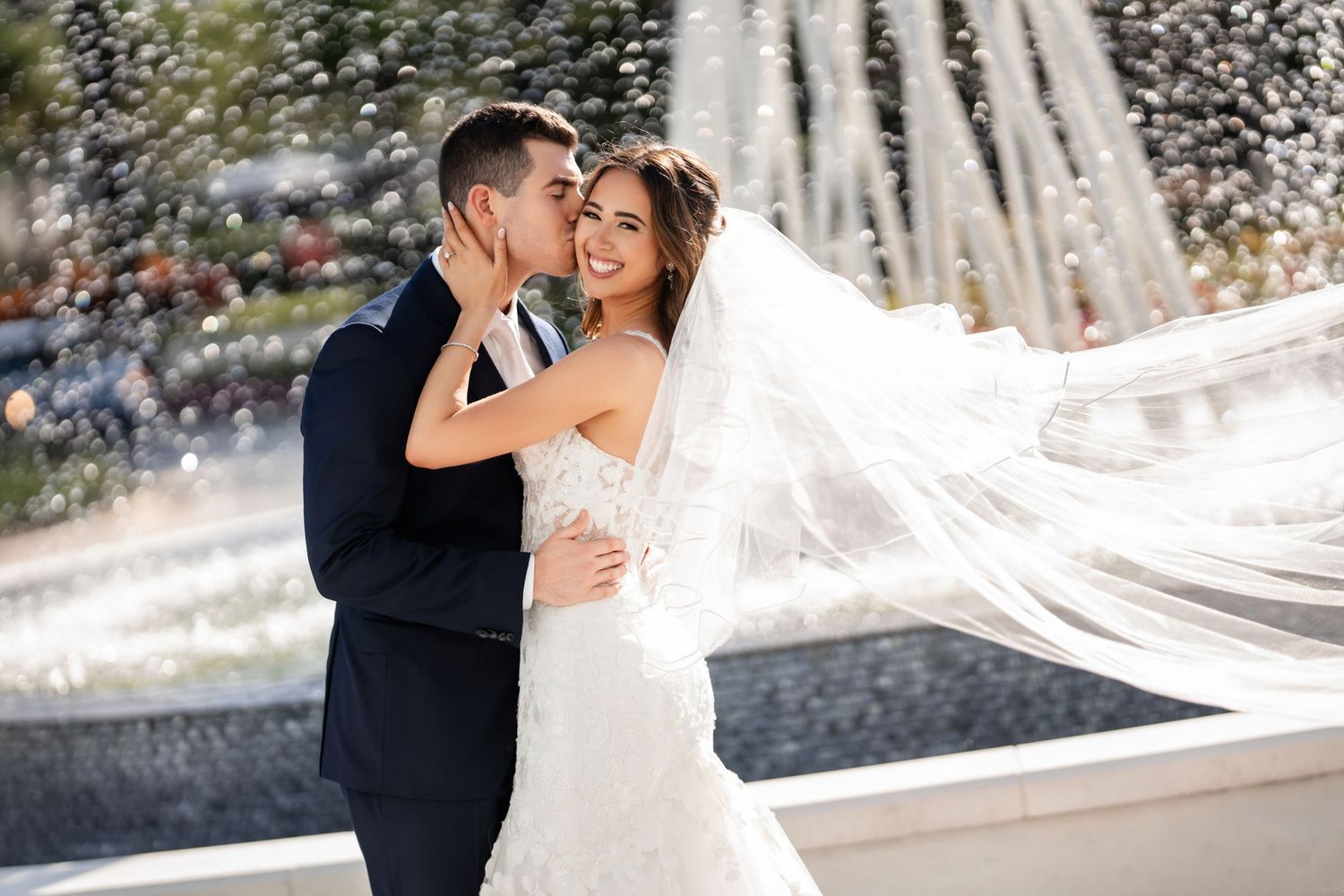 Bride and groom embracing outdoors with a flowing veil in front of a fountain—romantic wedding portrait.