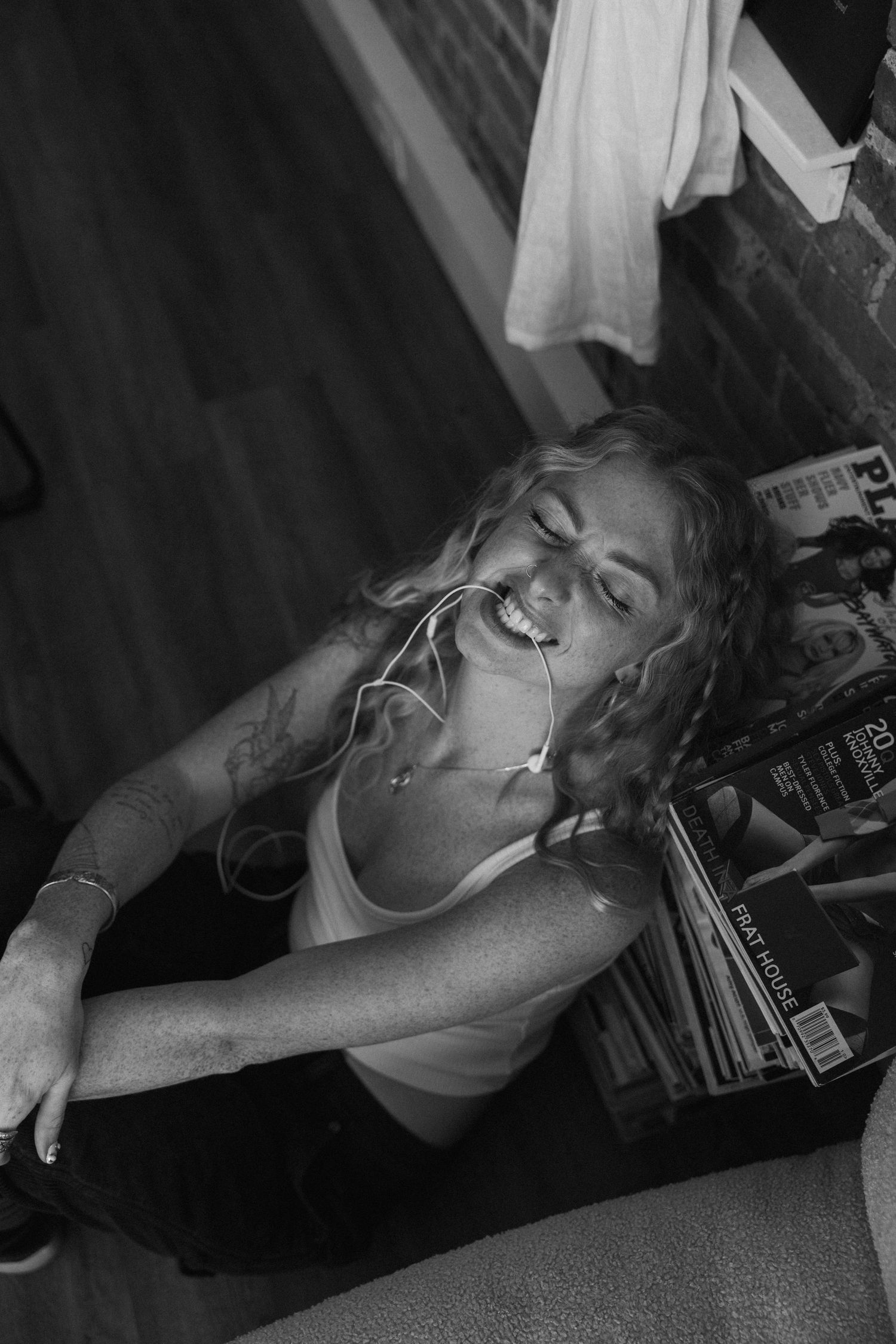 Black and white candid shot of laughing woman with earbuds sitting on floor next to stacked magazines.