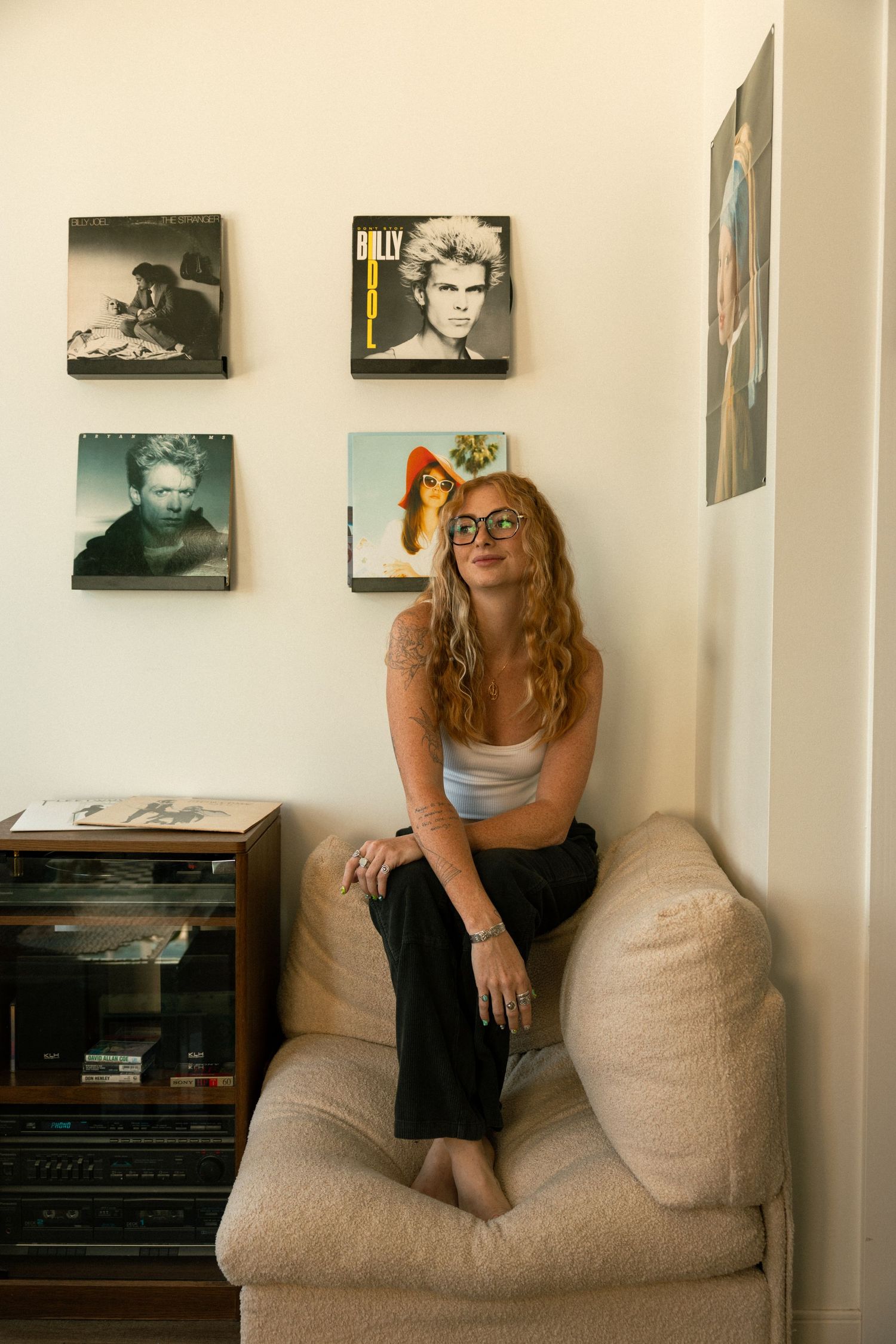 Young woman with glasses sitting in armchair beneath vintage Billy Idol and David Bowie album covers on wall.