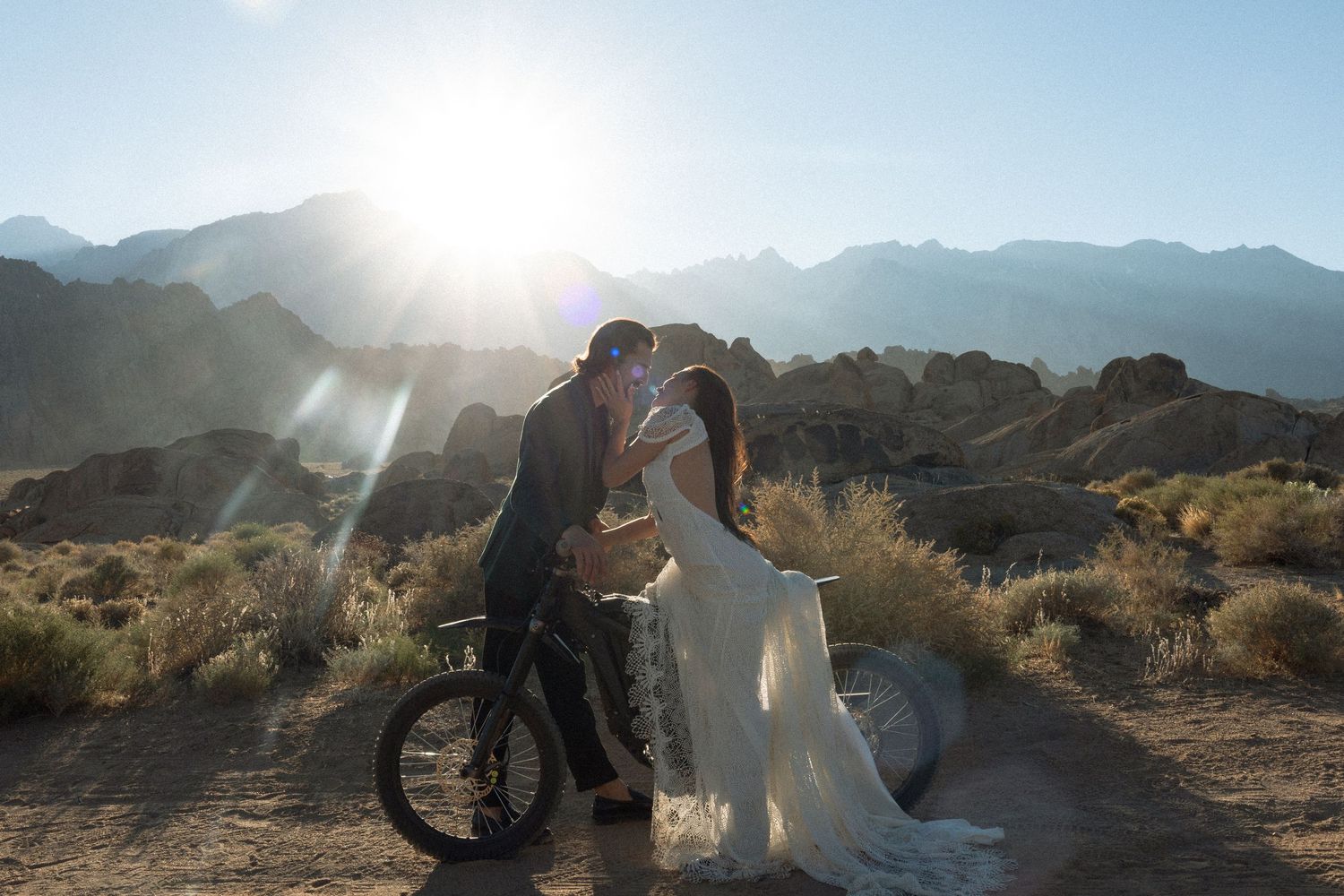 Bride and groom kissing at sunset in desert landscape with mountains, groom leaning on bicycle.