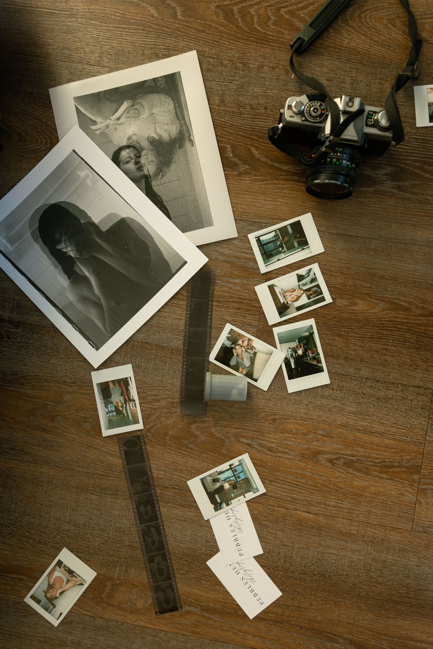 Vintage film camera, black and white prints, polaroid photos, and film strip arranged on wooden floor.