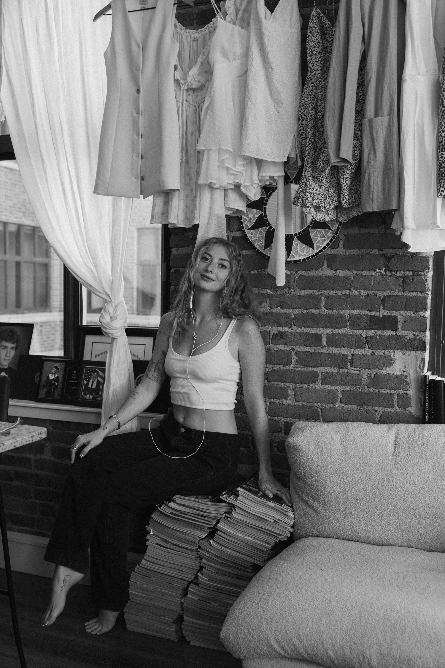 Black and white portrait of woman sitting on couch with stacked magazines beneath hanging vintage clothing rack.