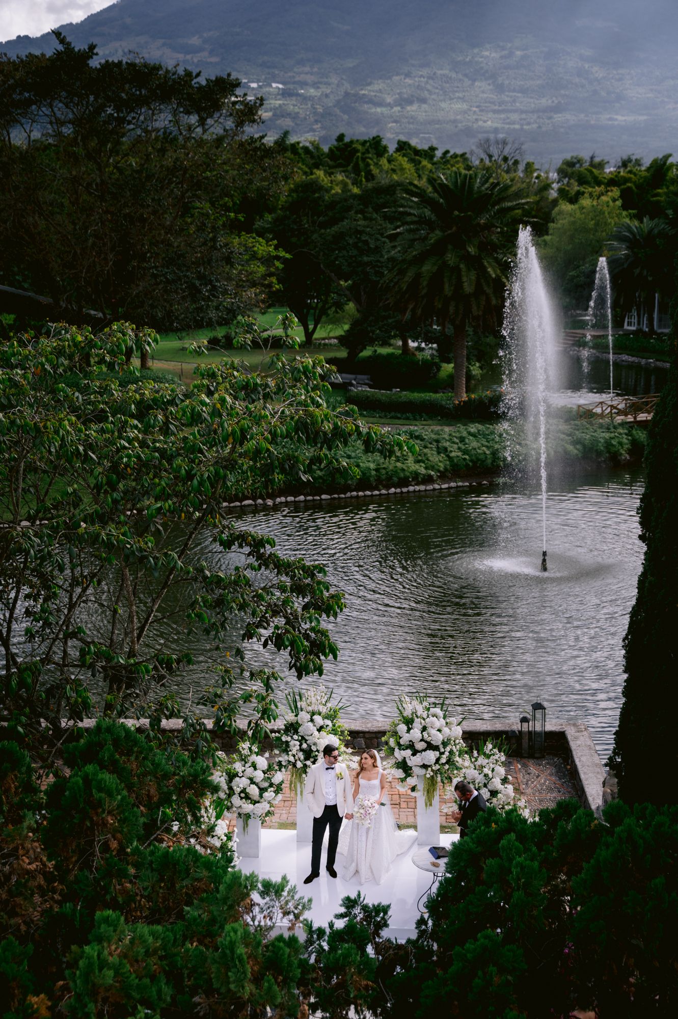 Aerial view of a wedding ceremony by a fountain in a lush garden setting with mountain backdrop.