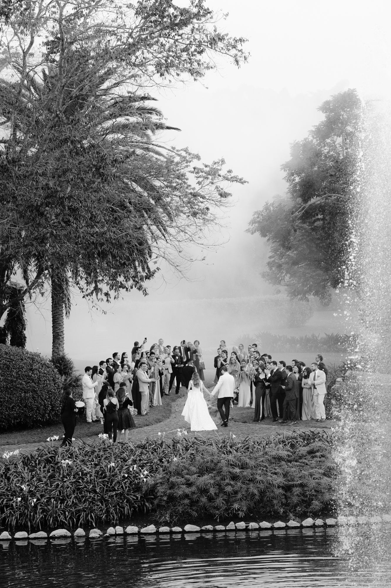 A vintage black and white photograph shows a wedding party gathered near a pond with palm trees in a misty setting.