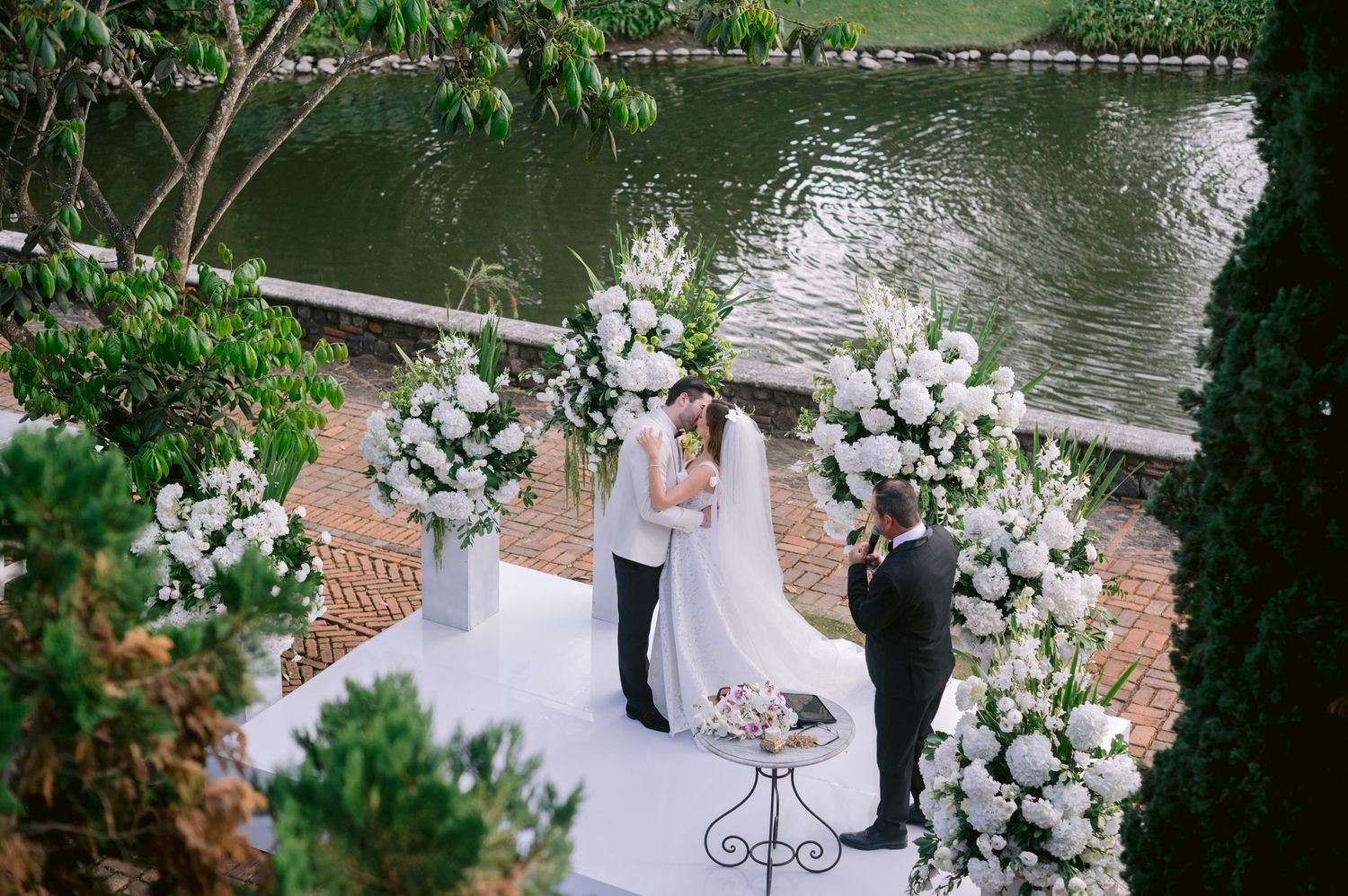 A romantic outdoor wedding ceremony by a tranquil lake with white floral arrangements and brick pathways.