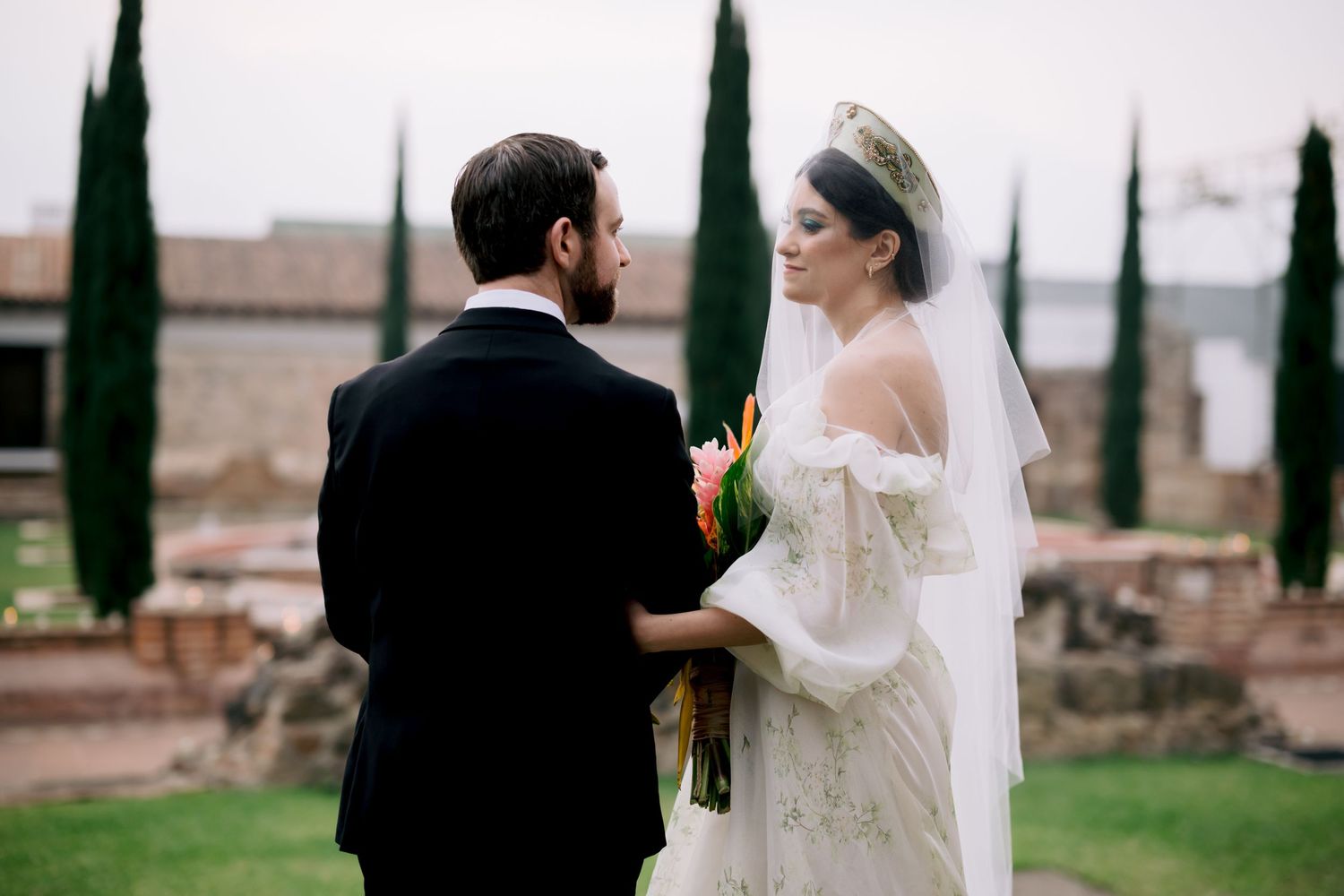 Bride and groom share an intimate moment among cypress trees at a historic stone venue.
