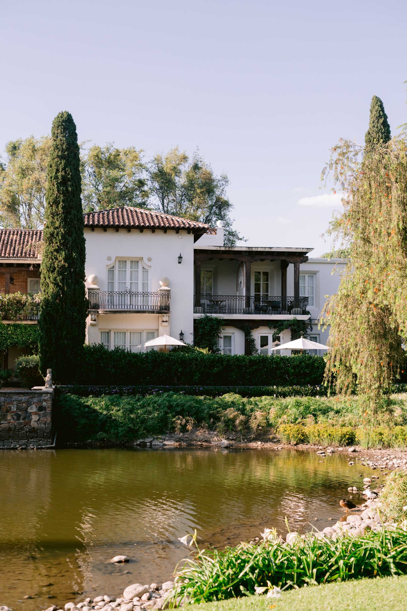 Mediterranean-style villa with balcony overlooking serene pond surrounded by cypress trees.