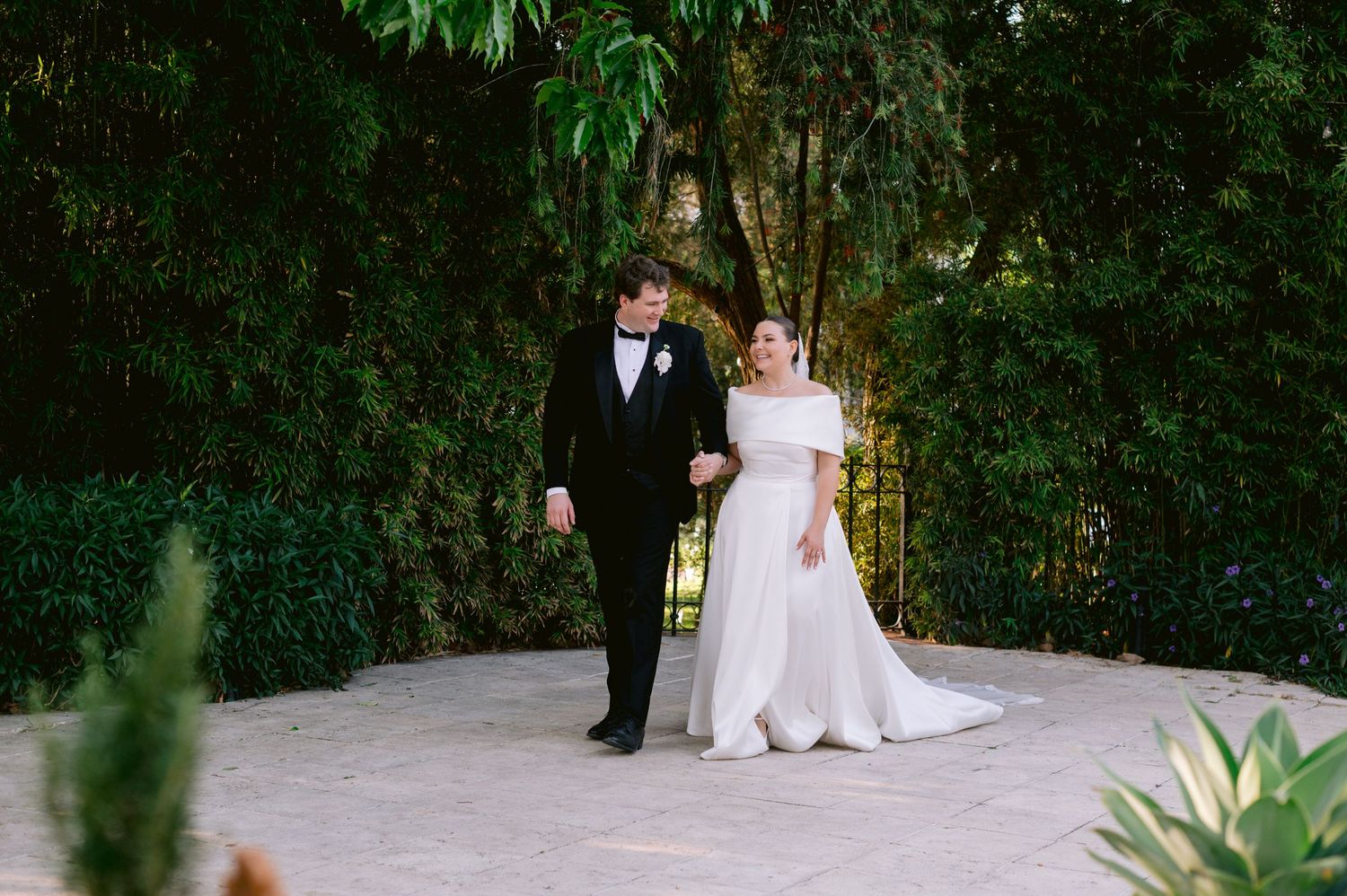 Wedding couple walking together on a garden path surrounded by lush greenery.