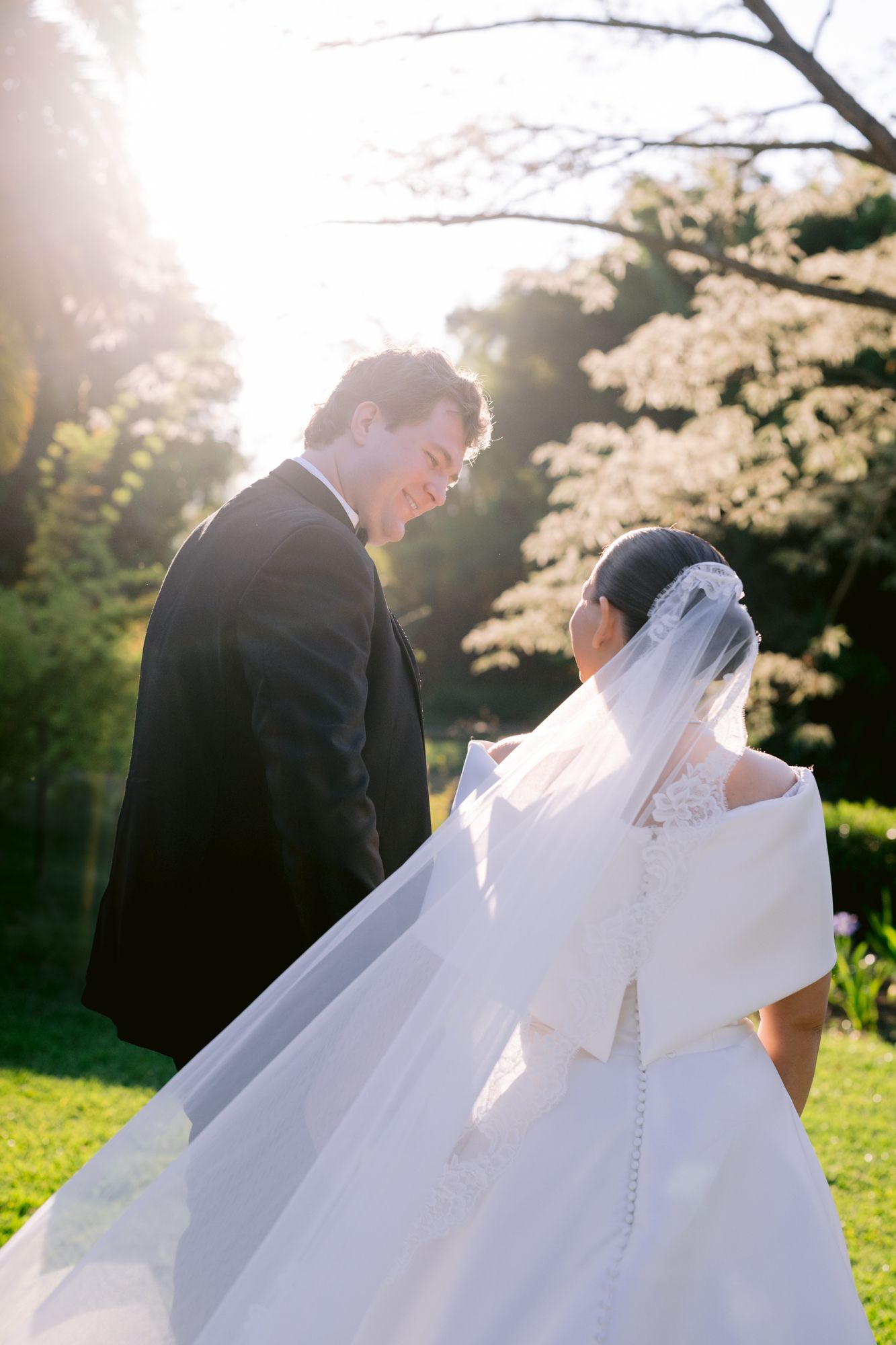 Sunlit garden wedding scene with flowing white dress and formal attire against blooming trees.