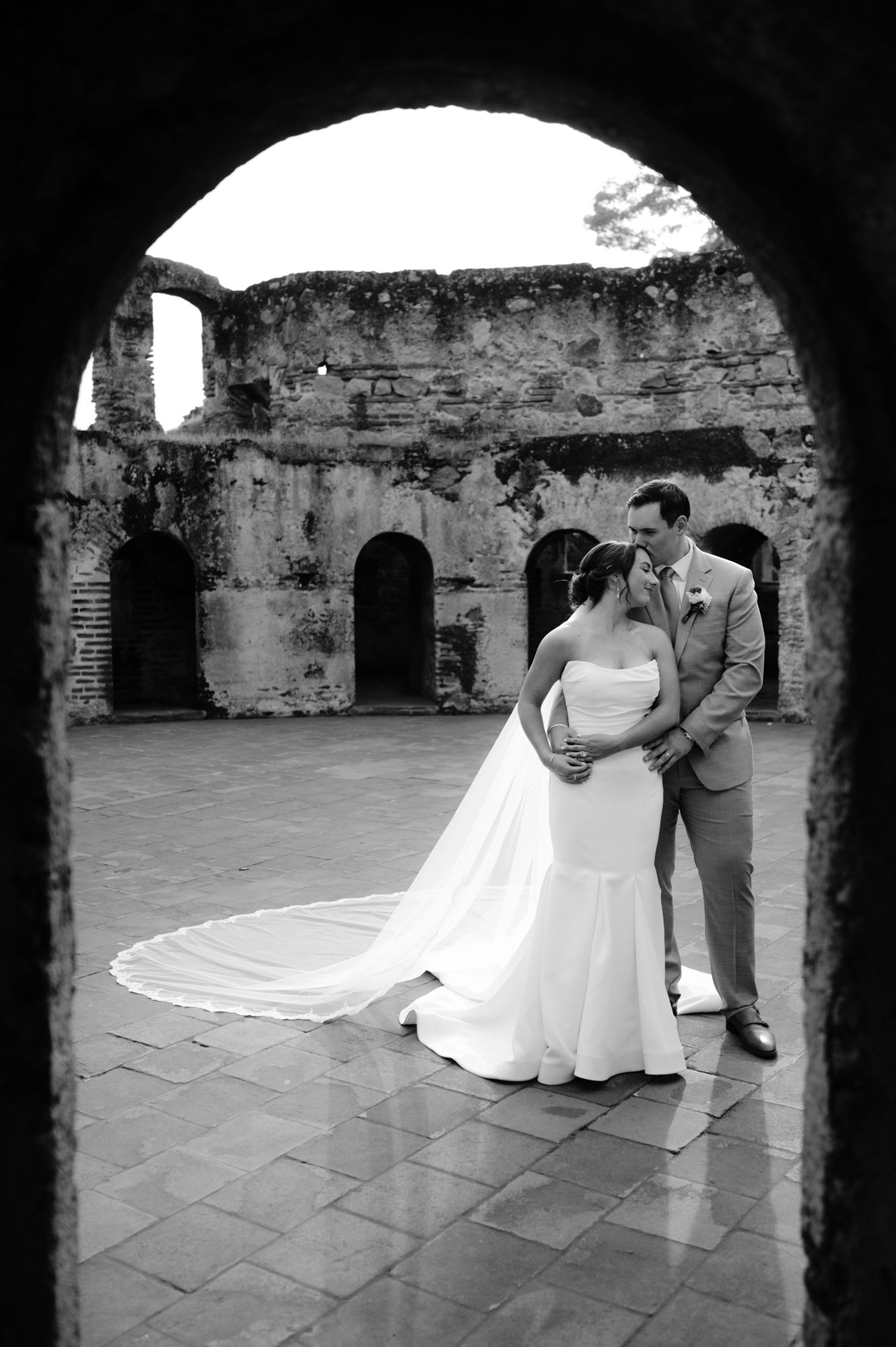 Black and white wedding photo captured through stone archway showing couple in elegant embrace.