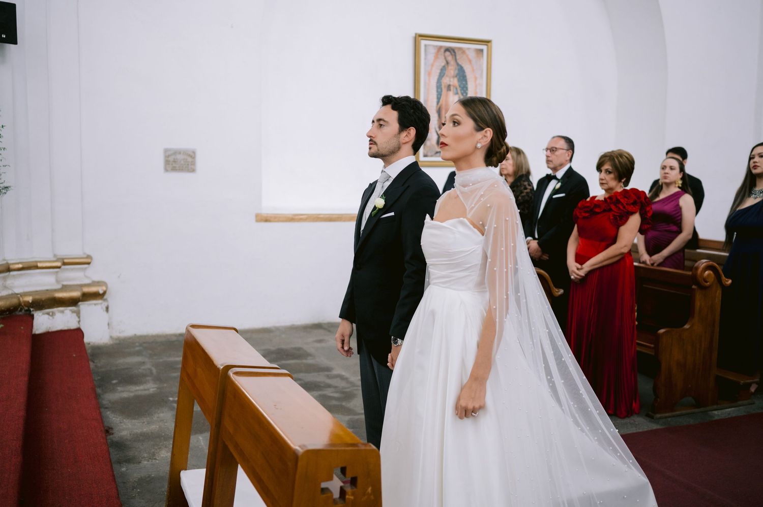 Bride in flowing white dress and groom in dark suit stand together during church ceremony.