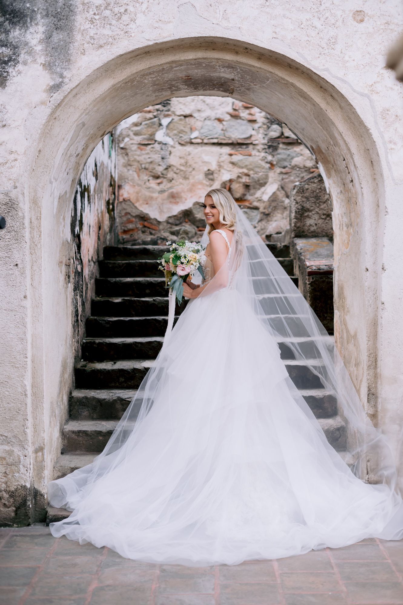 A bride in a flowing white ball gown and veil stands under a stone archway near weathered steps.