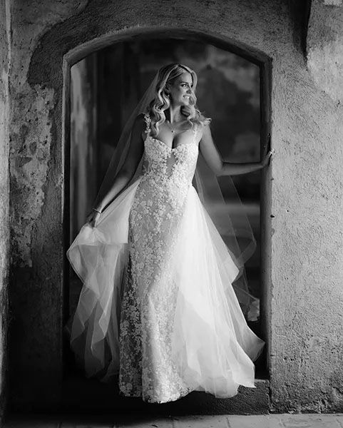 Elegant bride in a lace fitted wedding gown with flowing tulle overlay stands in stone archway in black and white photo.