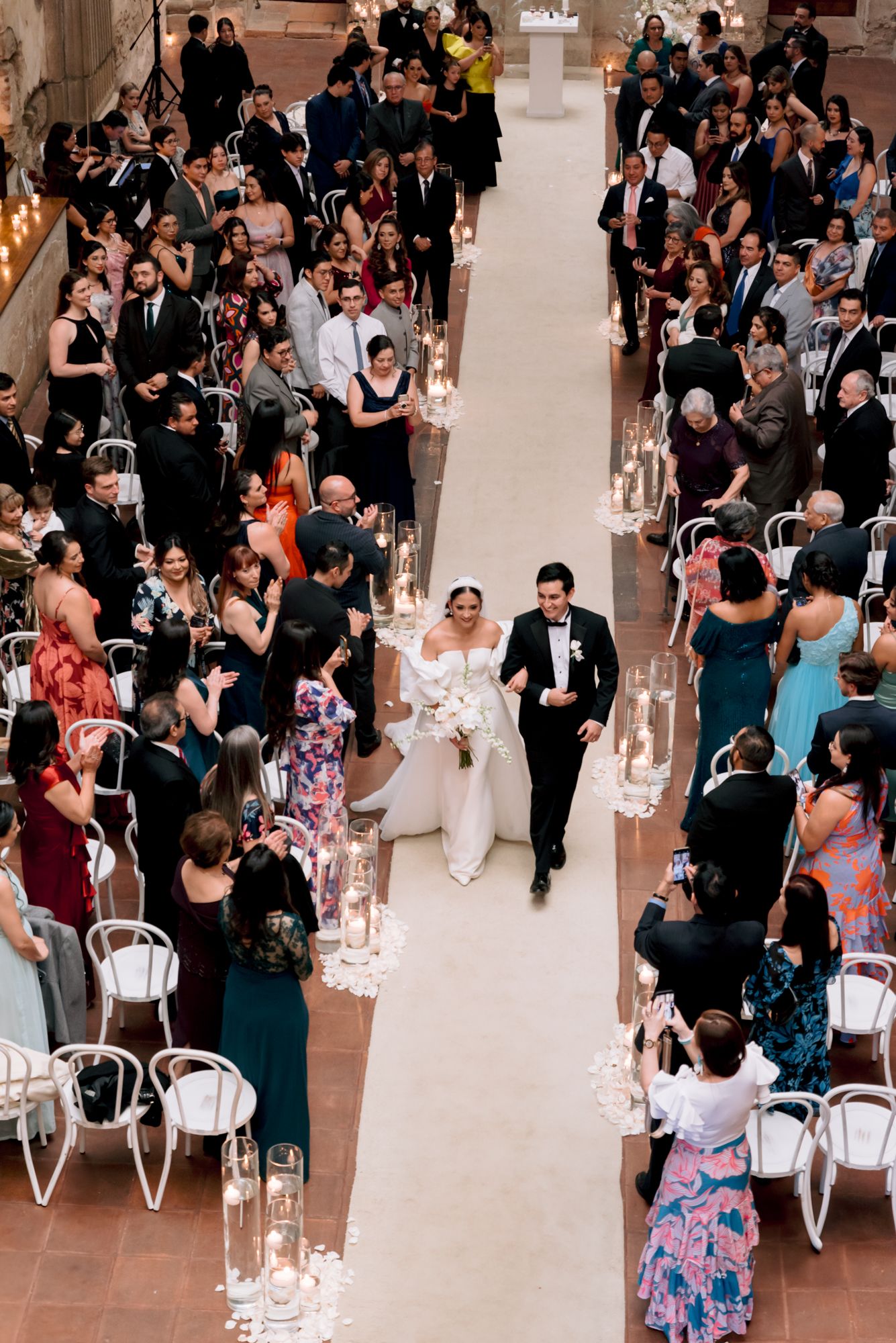 A couple walks down the aisle at their wedding ceremony while guests stand and watch from white chairs along both sides.