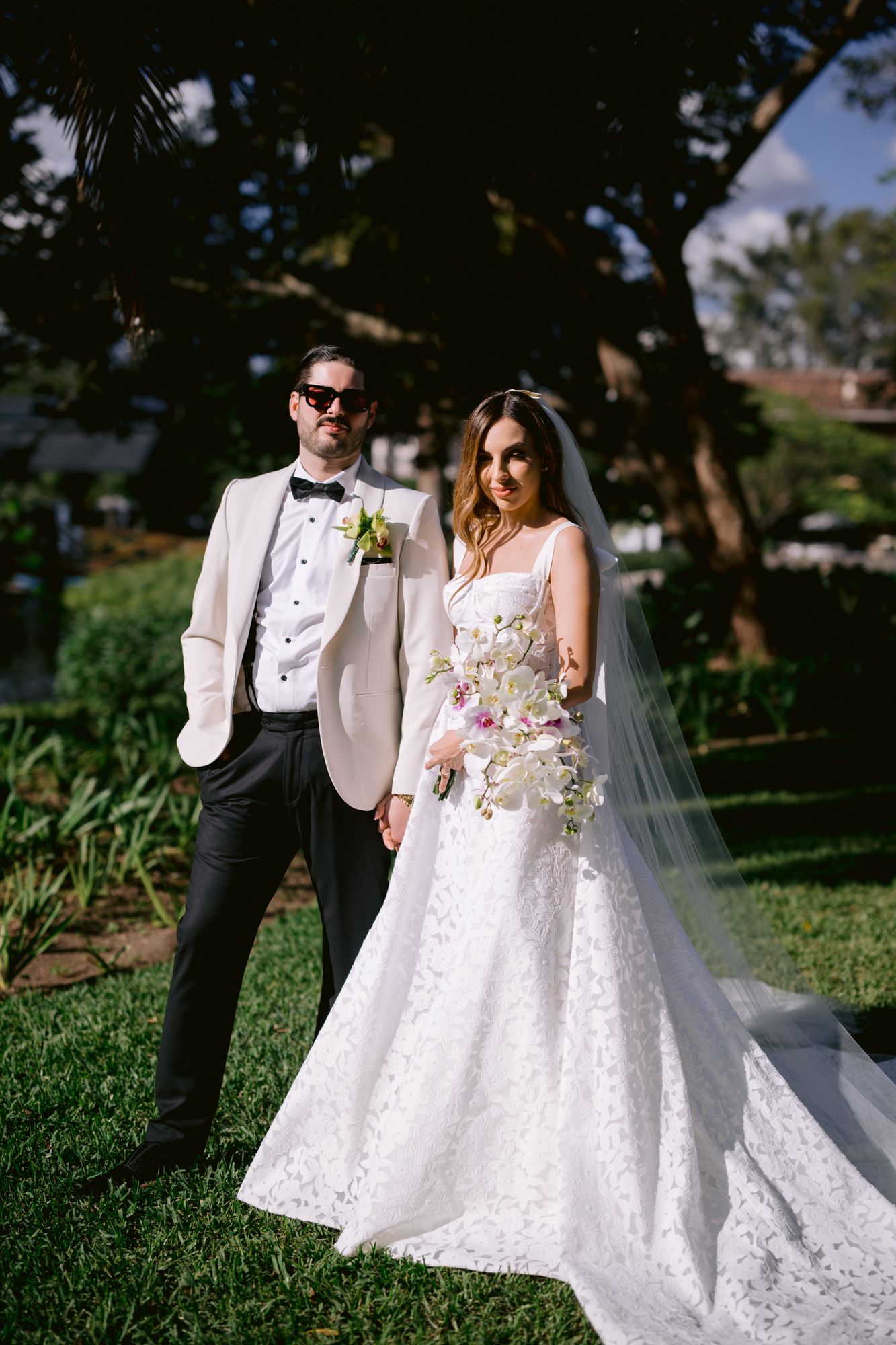 A couple poses together in wedding attire with a white lace gown and white tuxedo jacket in an outdoor garden setting.